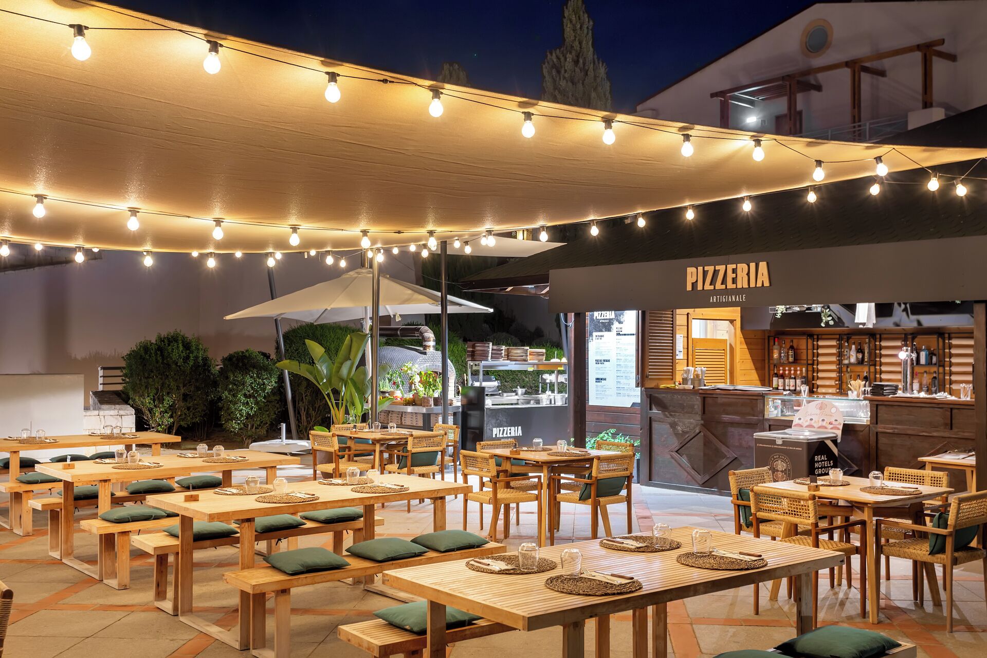An outdoor dining area with string lights, wooden tables, and green cushions.