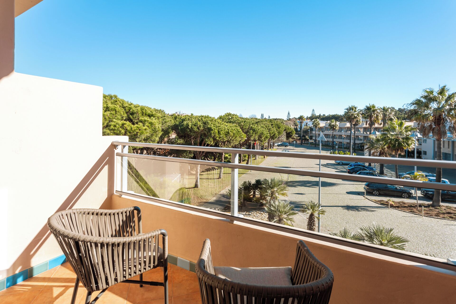 A balcony with wicker chairs, a clear view of a parking area, and a backdrop of trees and buildings.