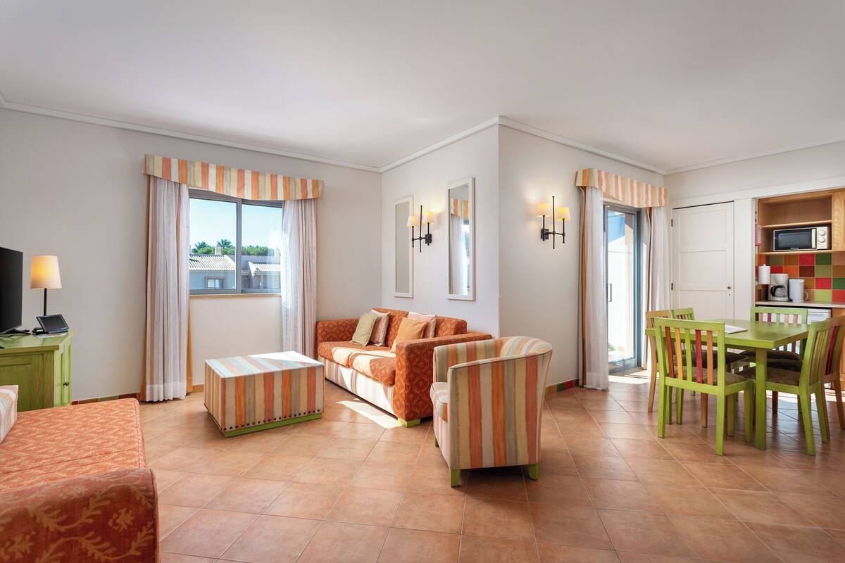 A living room with a sofa, armchairs, a coffee table, and a TV. There is a dining area with a table and chairs.