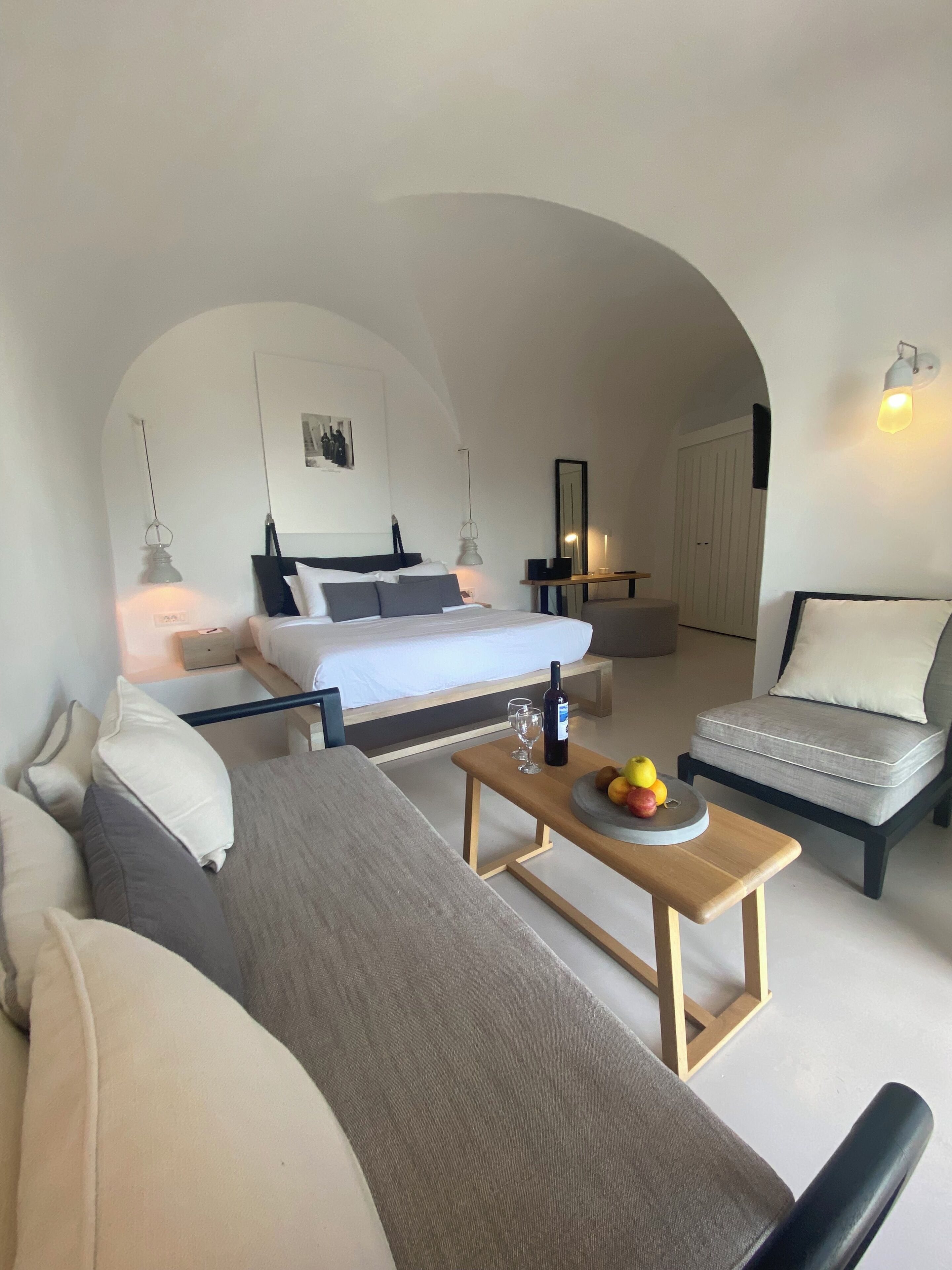 Une chambre d’hôtel moderne avec un lit, un canapé, une petite table avec des fruits, un miroir et une porte.