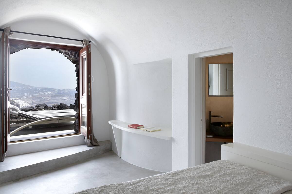 Ein Schlafzimmer mit einer gewölbten weißen Wand, einem Fenster mit Bergblick, einem Nachttisch mit einem Buch und einem Badezimmer mit Waschbecken und Spiegel.