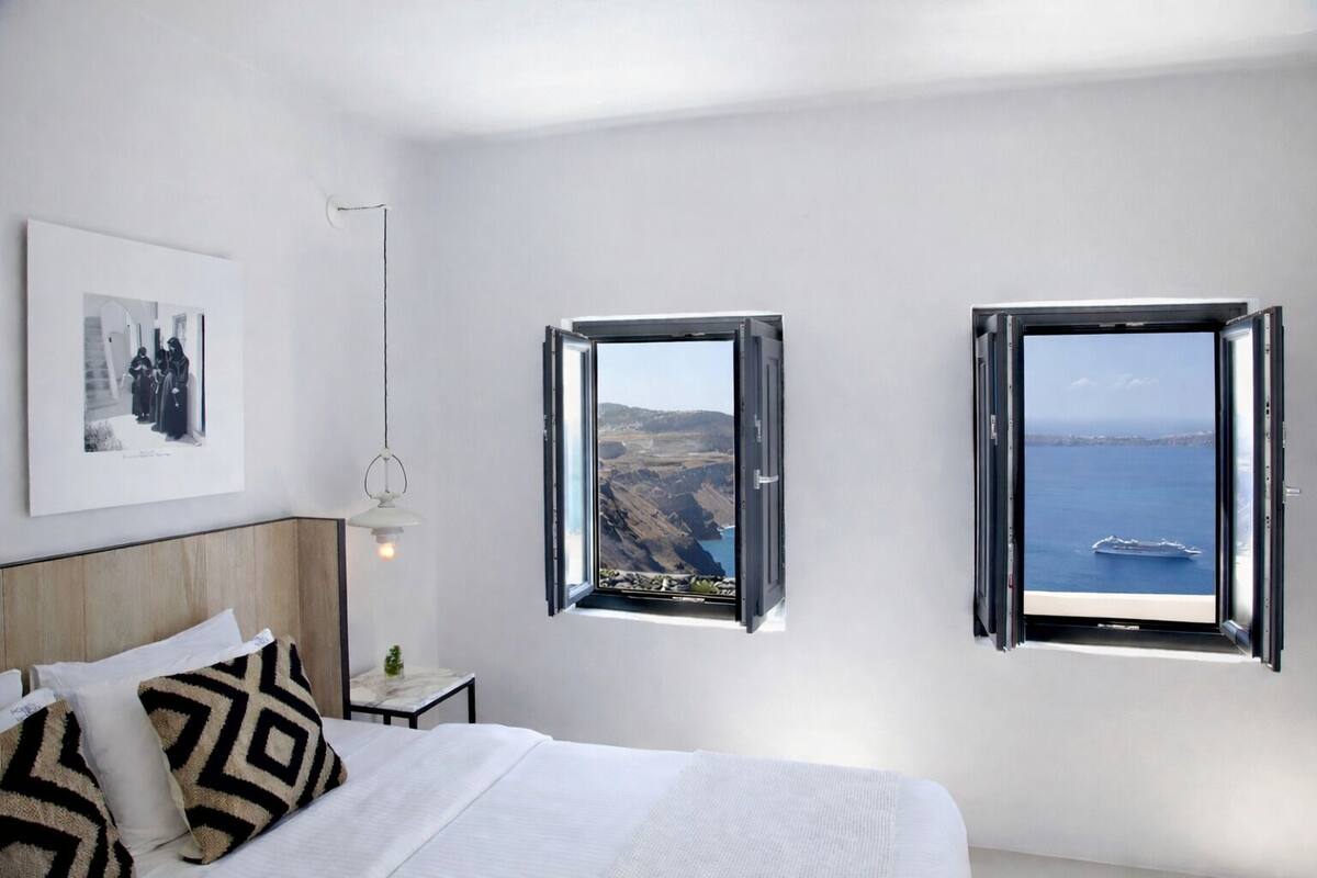 Ein Schlafzimmer mit Bett, Nachttisch, Pendelleuchte und zwei Fenstern mit Blick auf eine Küstenlandschaft.