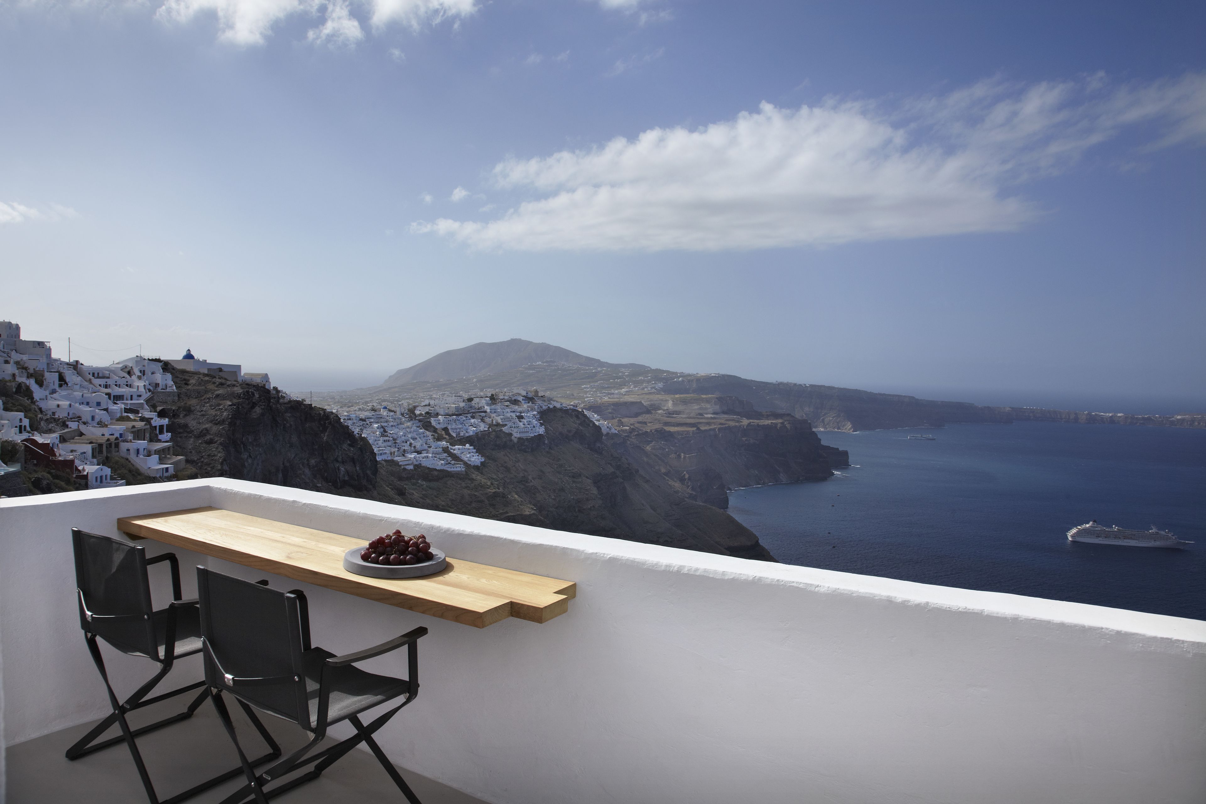 A balcony with a wooden table and two chairs, overlooking a coastal landscape with white buildings and a ship in the distance.