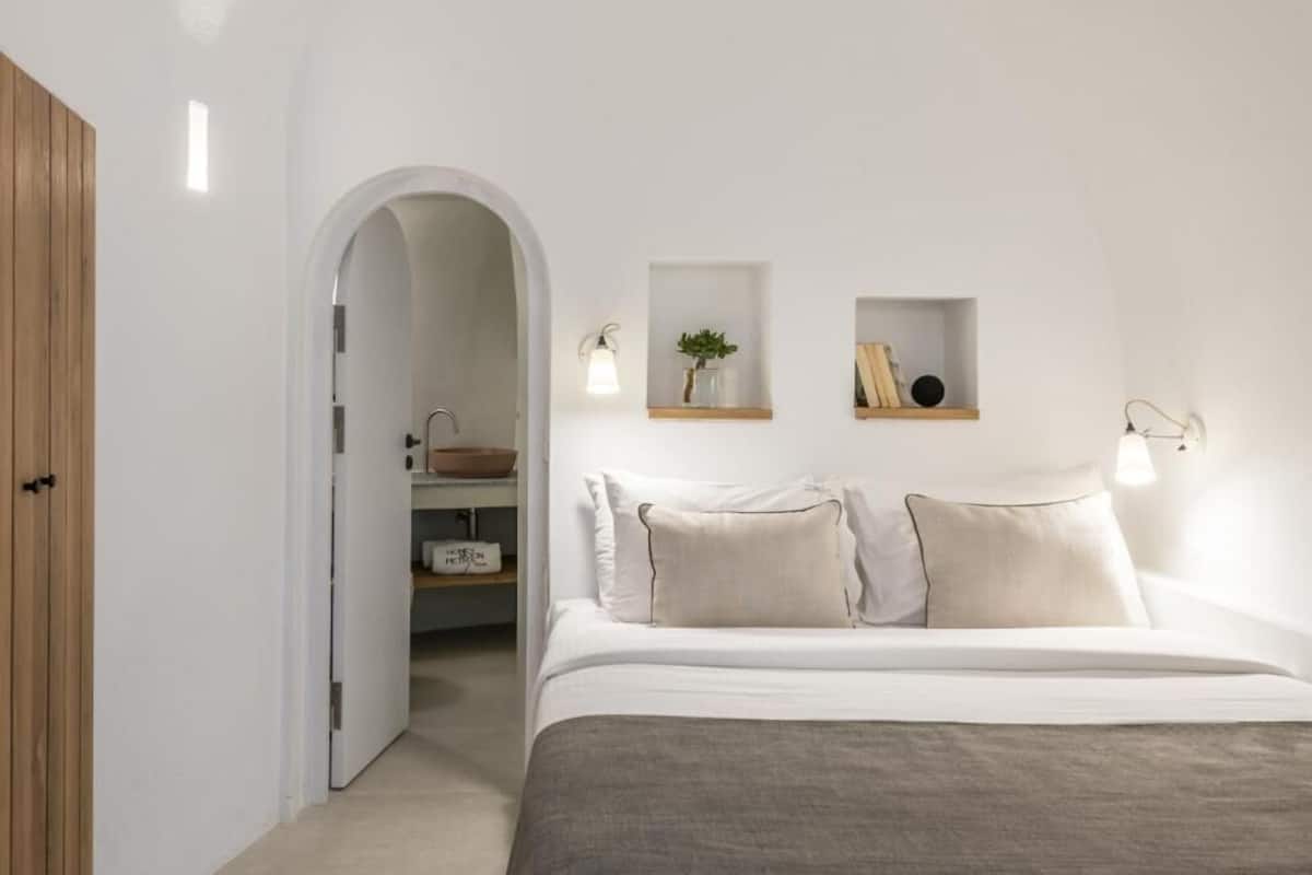 Ein Schlafzimmer mit Bett, einem Holzschrank, einer weißen Wand mit Regalen und einem zur Badezimmertührenden Durchgang.