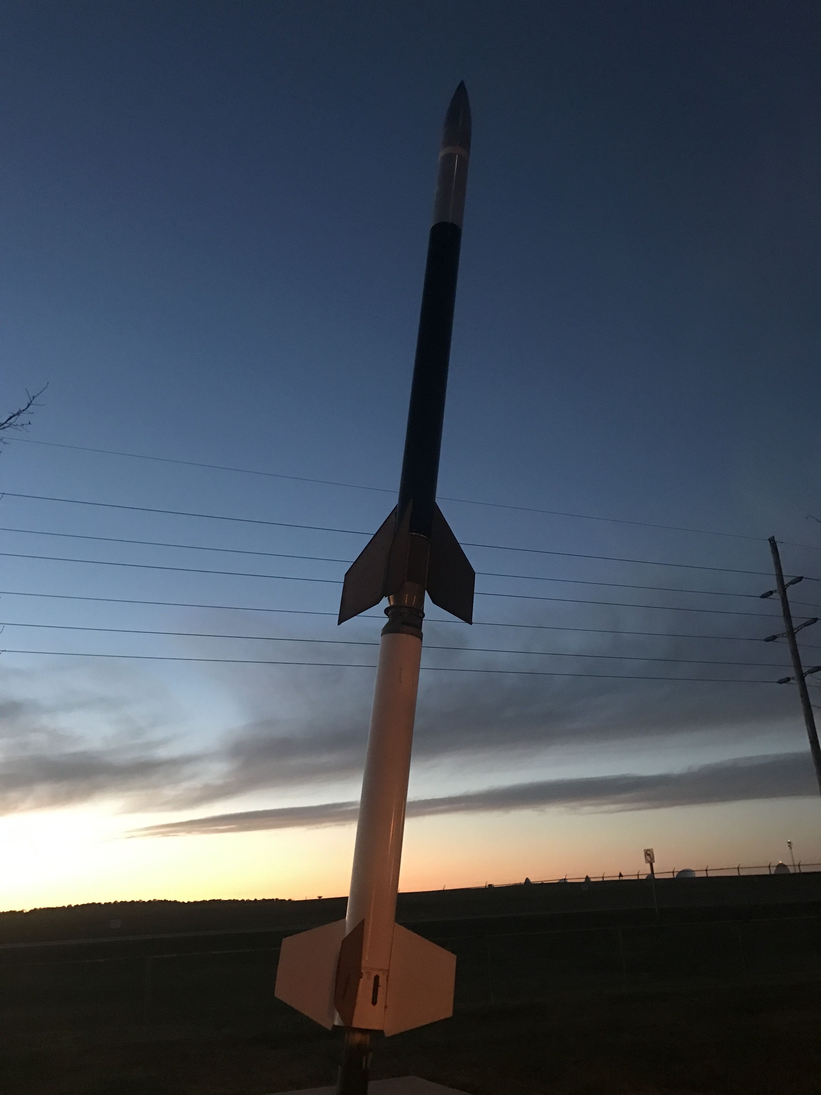 Un cohete vertical en un campo, con un cielo despejado de fondo.