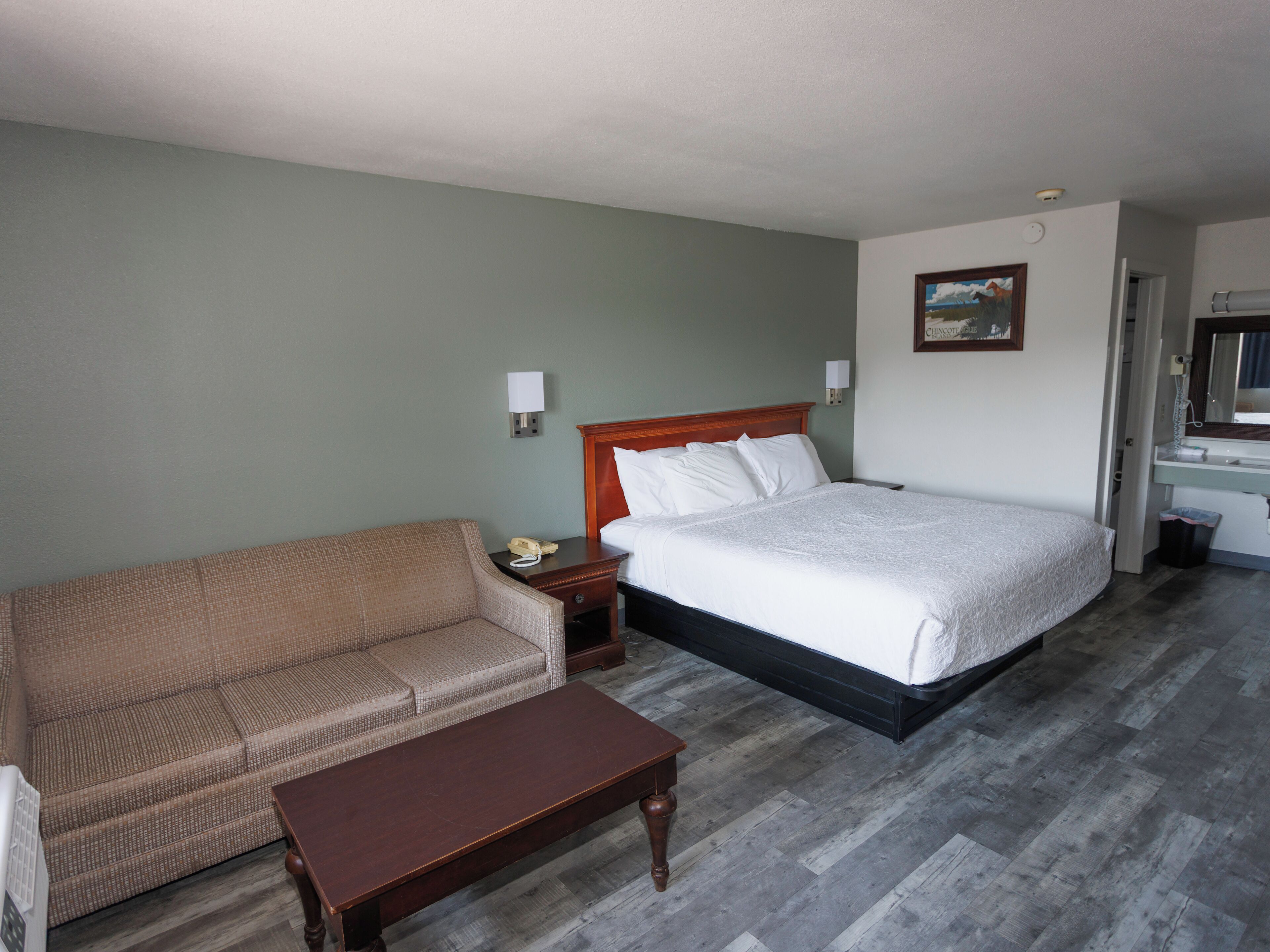 Habitación de hotel con cama, sofá, mesita pequeña, mesita de noche, lámpara de pared y un cuadro.