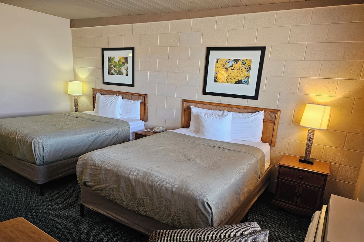 Habitación de hotel con dos camas, cabecera de madera, mesita de noche con lámpara, una pequeña mesa y una silla.