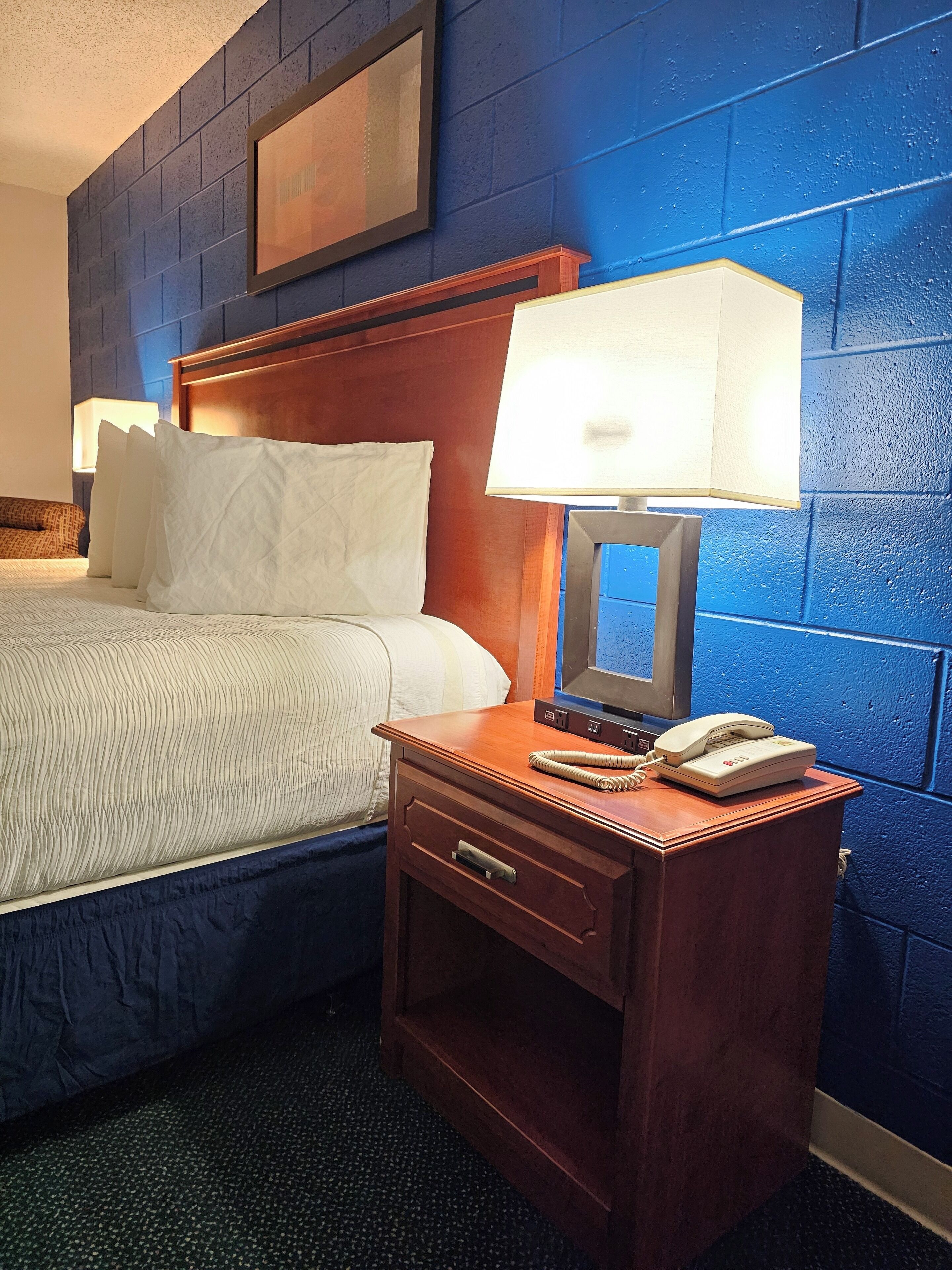 Habitación de hotel con cabecera de madera, cama con ropa de cama blanca, mesita de noche con lámpara y teléfono, y pared de ladrillo azul.