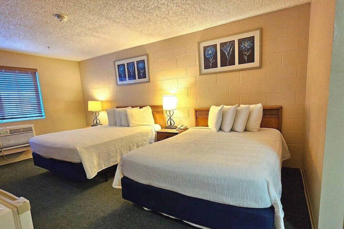 A hotel room with two beds, a window with blinds, a heater, and framed pictures on the wall.