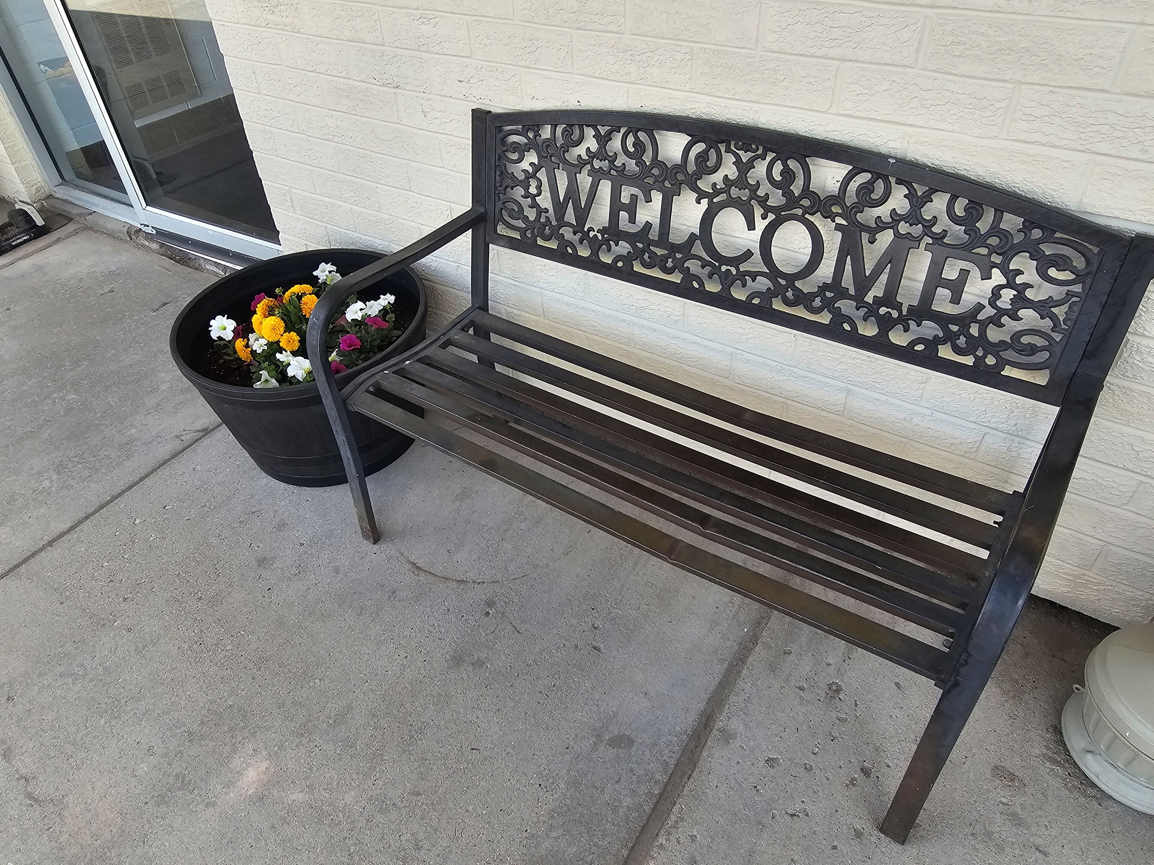 A black metal bench with a 'WELCOME' sign, placed against a white brick wall.