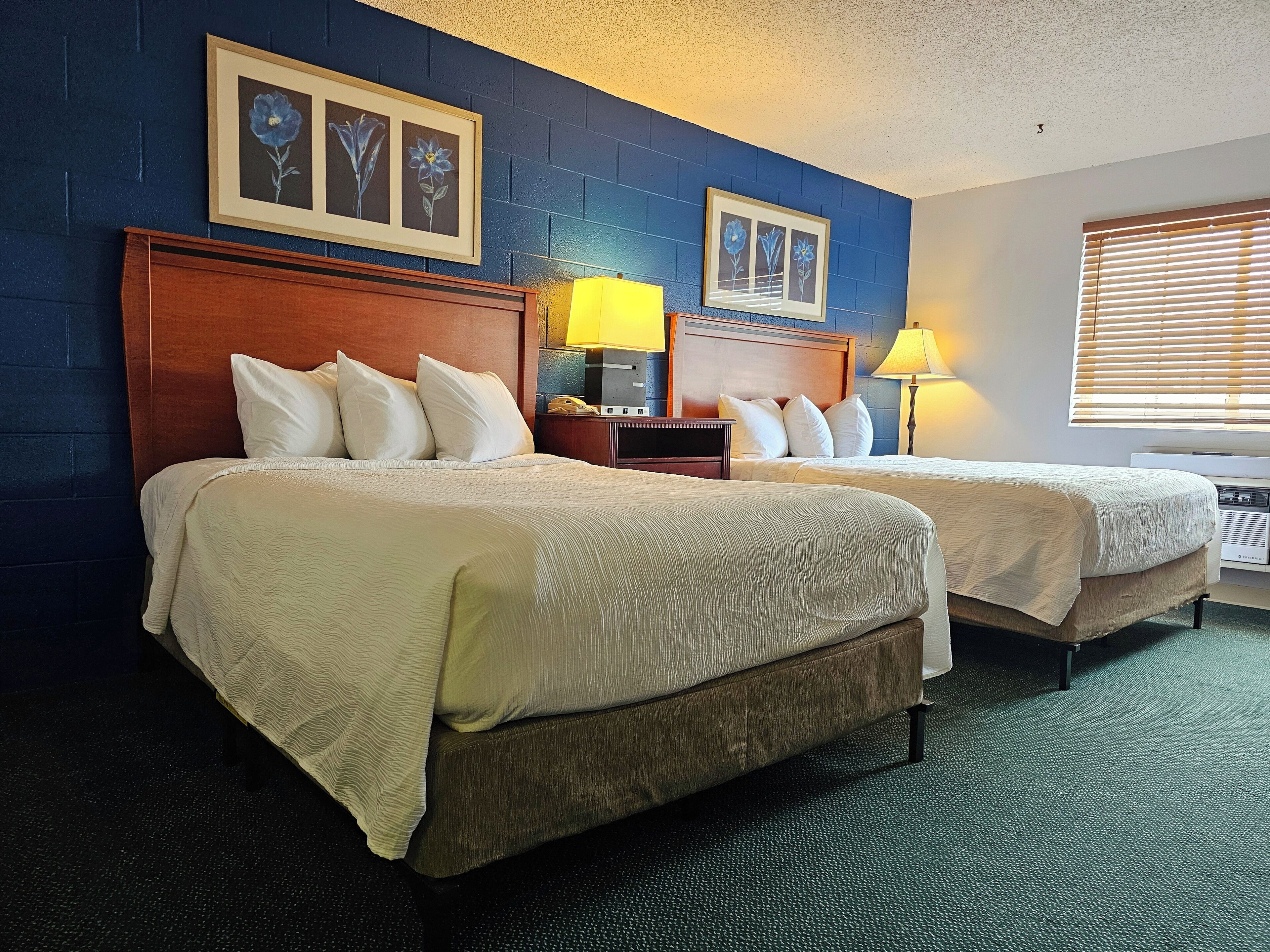 A hotel room with two beds, a wooden headboard, a nightstand with a lamp, and framed floral artwork on the wall.