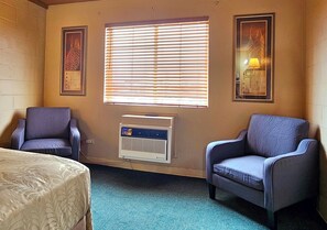 A room with two armchairs, a bed, a window with blinds, and a wall-mounted heater. - 1st Choice Inn Rawlins (Rawlins)