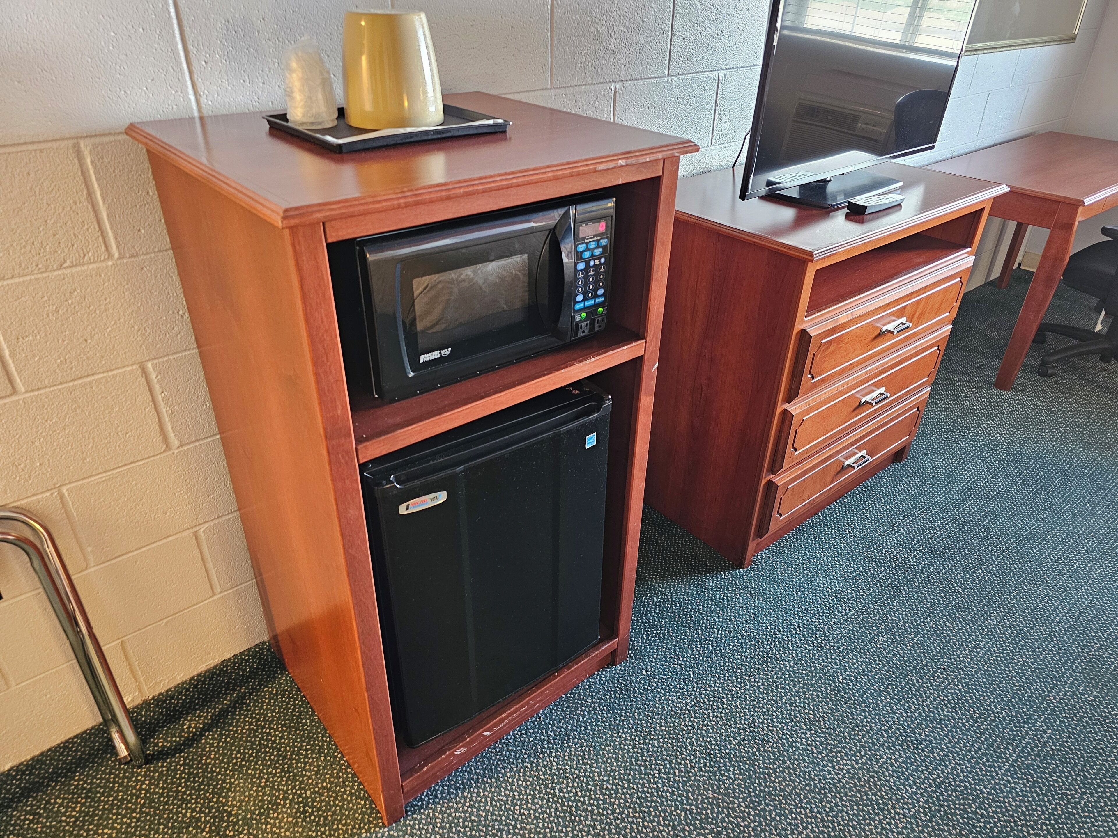 A compact kitchenette with a microwave and refrigerator, a wooden desk with drawers, and a television on a stand.