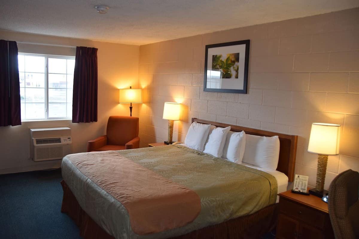 Habitación de hotel con cama, dos lámparas de noche, una silla, ventana con cortinas, aire acondicionado y un cuadro en la pared.