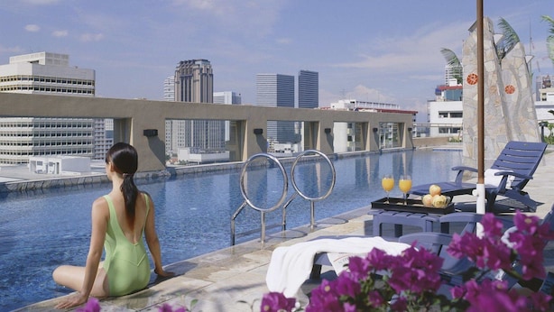 Una donna seduta a bordo piscina con vista sulla città, sedie a sdraio e un muro di pietra con fiori.