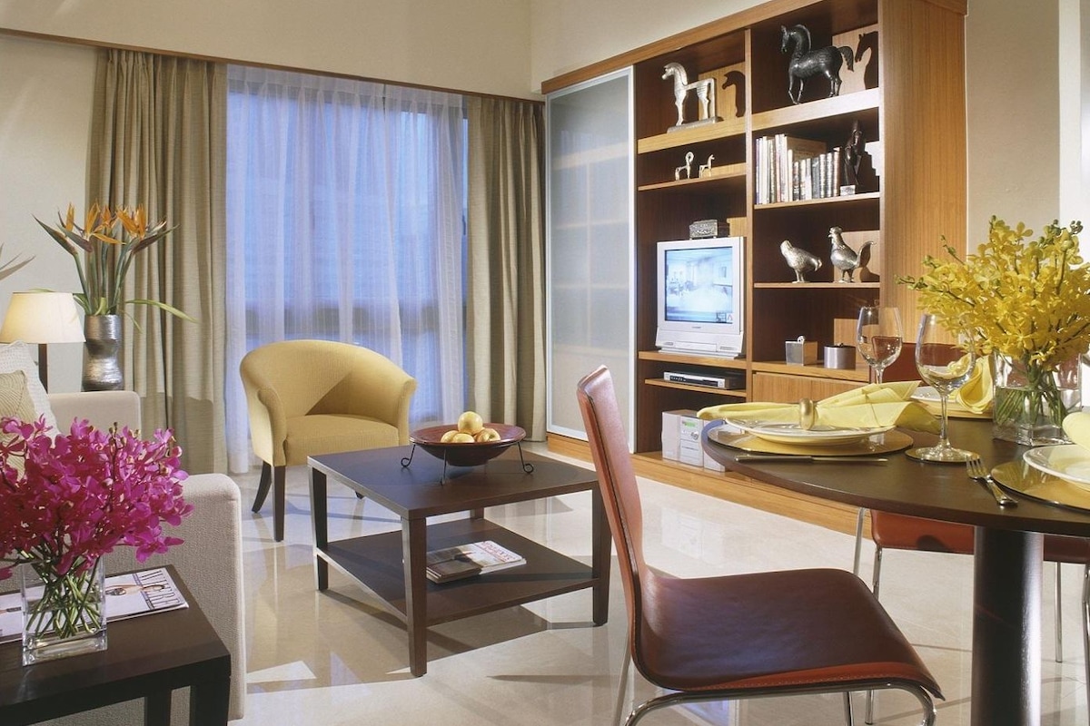 Una habitación de hotel moderna con zona de comedor, un sofá cómodo, televisión y estantería con objetos decorativos.