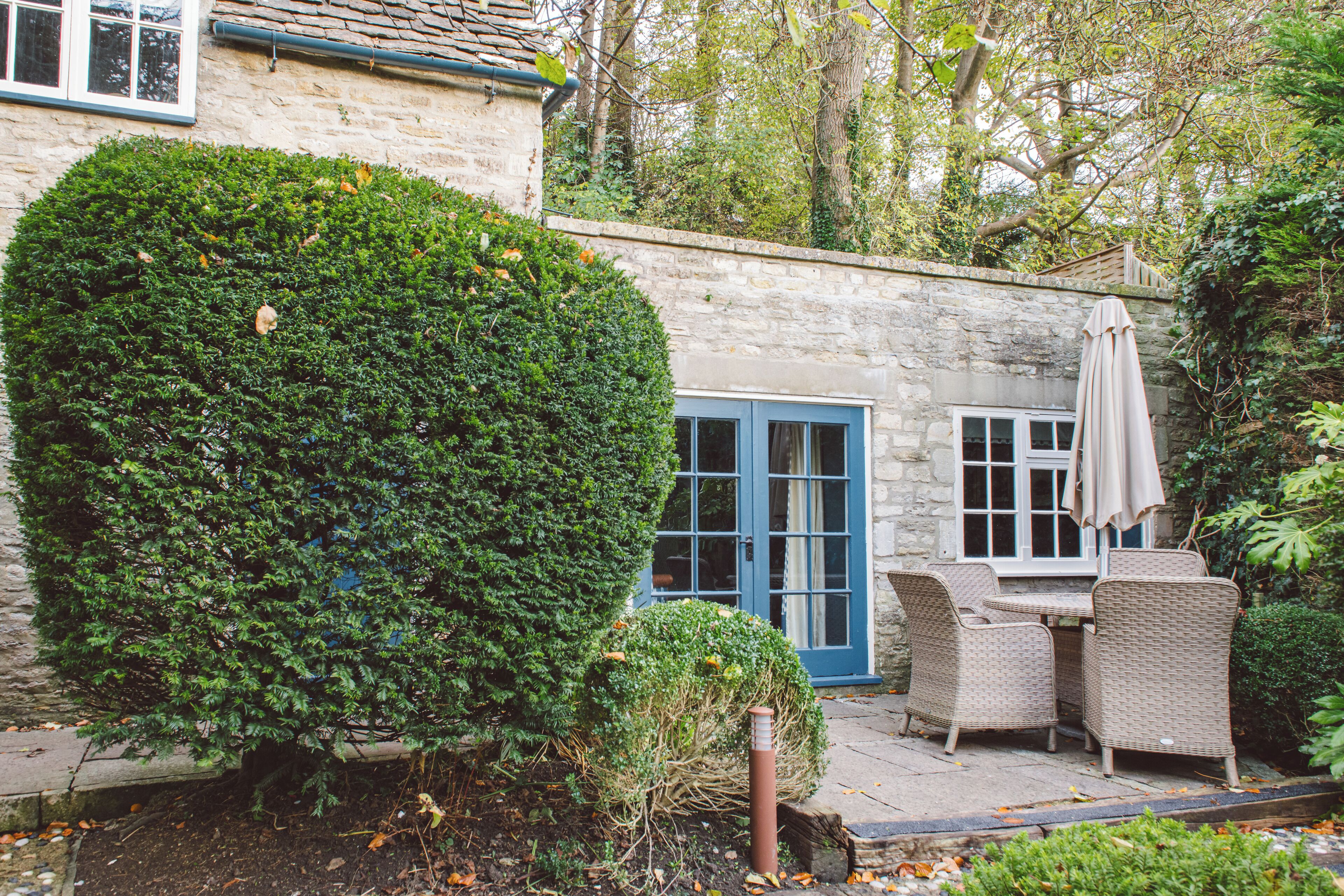 A stone building with a blue door and windows, surrounded by greenery and a patio with wicker furniture.