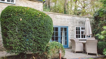 A stone building with a blue door and windows, surrounded by greenery and a patio with wicker furniture.