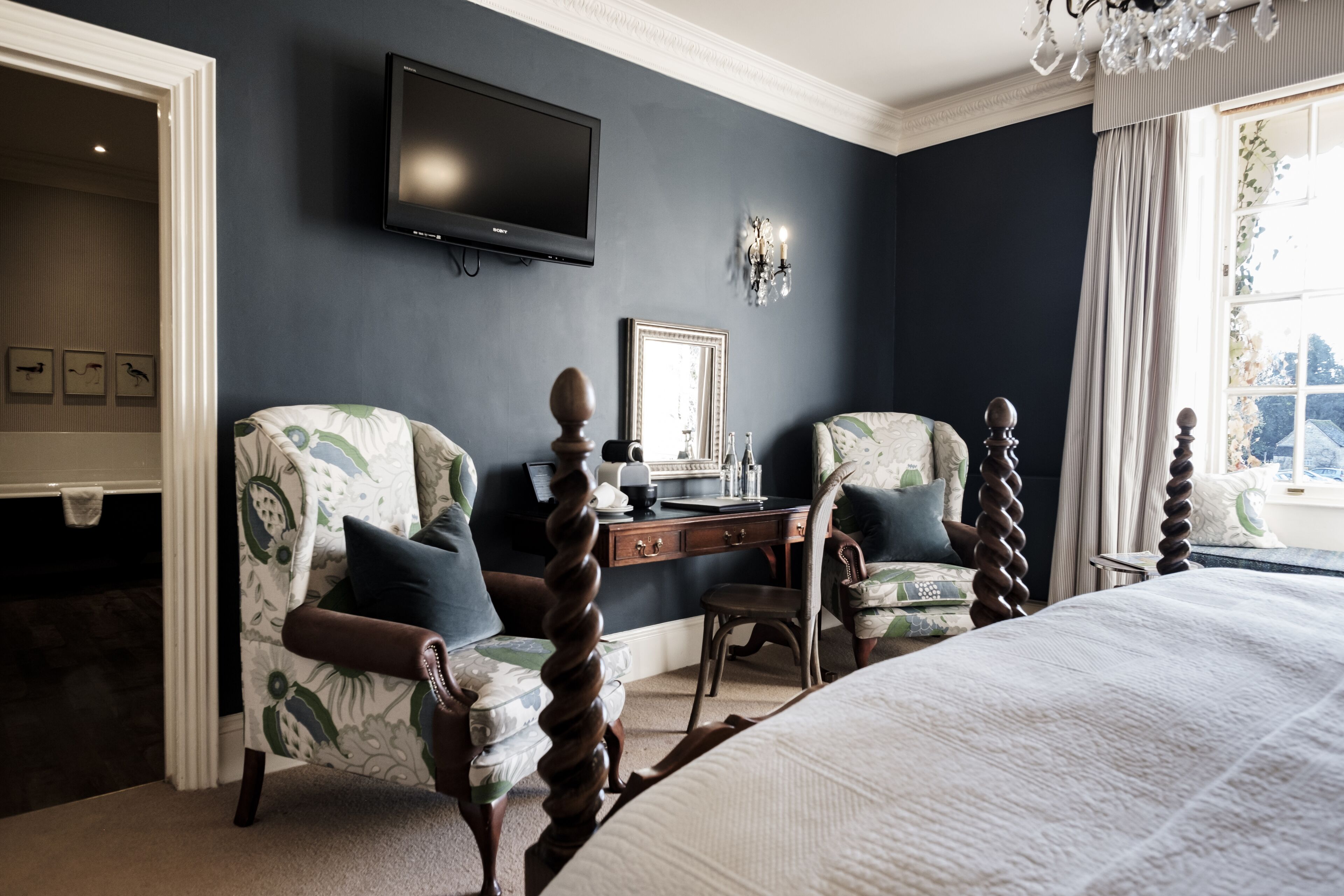 A bedroom with a large bed, two armchairs, a desk, and a television on the wall.