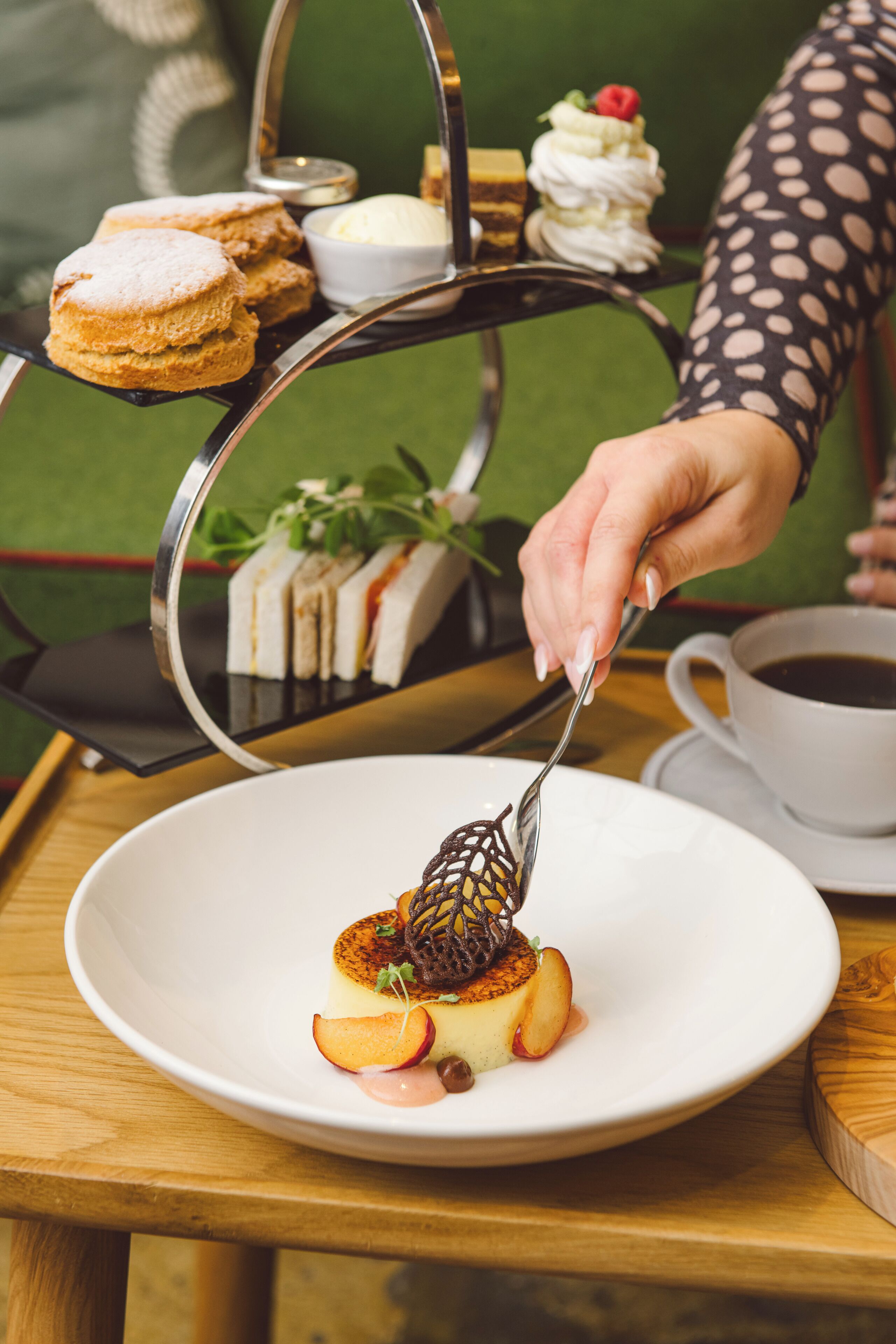 Un plato de postre con un pastel, fruta y salsa, una taza de café, y una bandeja con pasteles y crema.