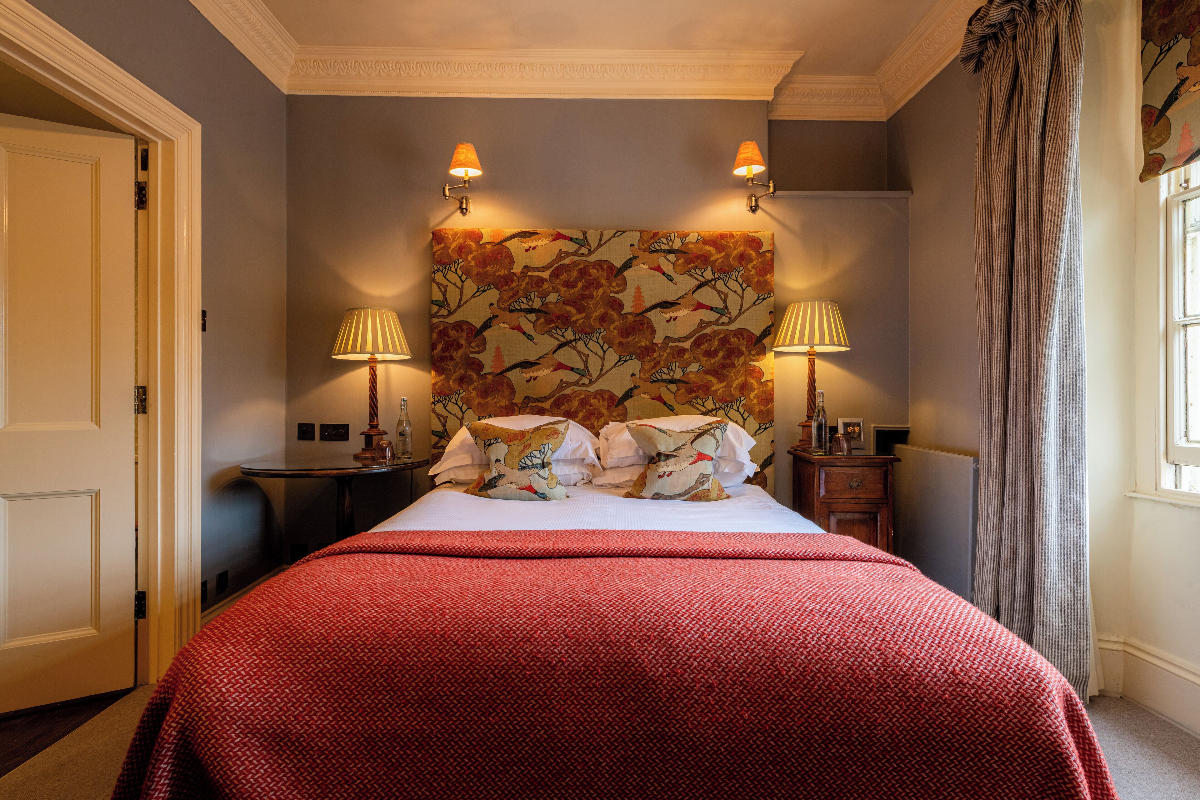 A neatly made bed with a red bedspread, two wall-mounted lamps, and a floral headboard.