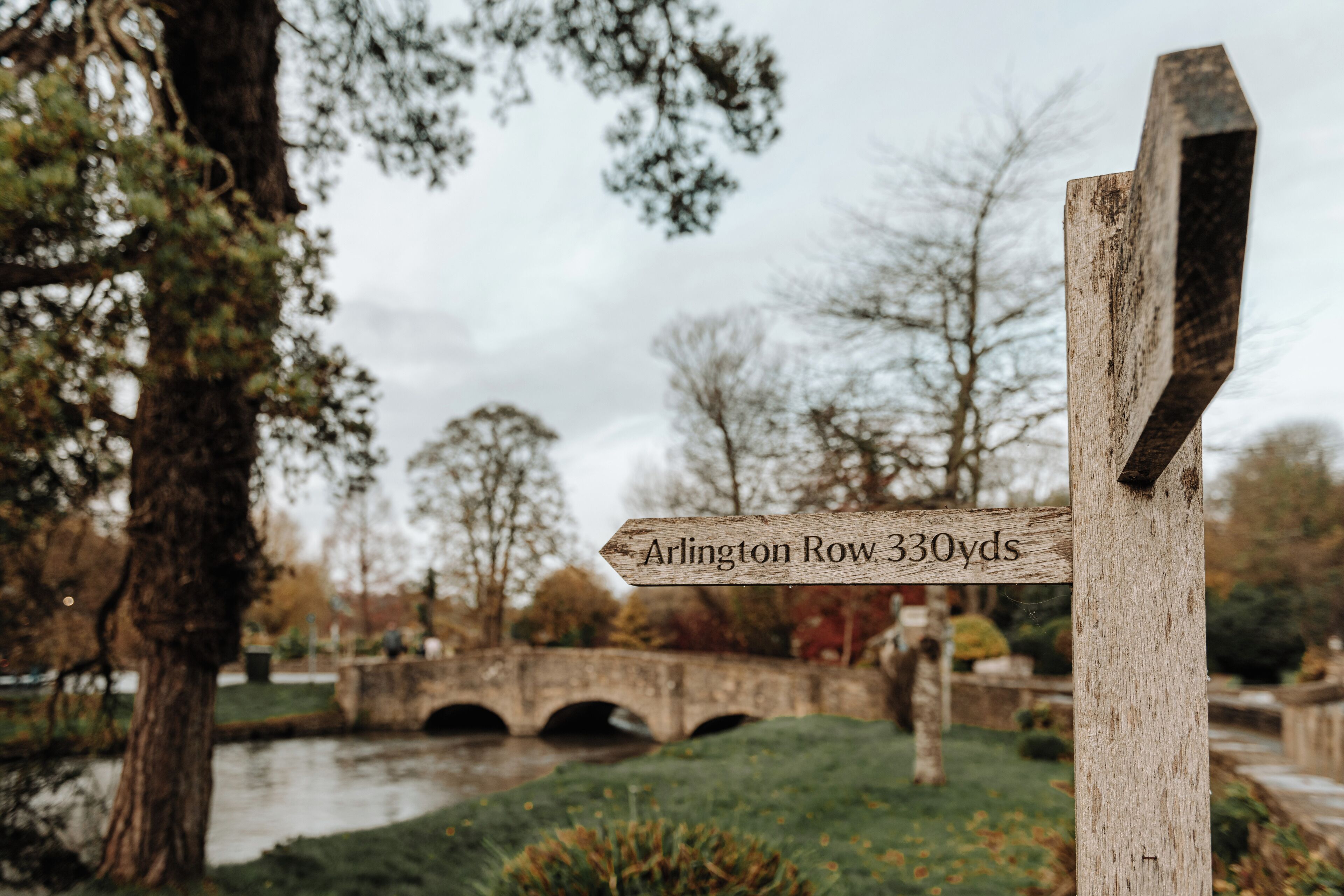 A wooden signpost with 'Arlington Row 330yds' written on it, set against a backdrop of a stone bridge over a river, surrounded by trees and greenery.