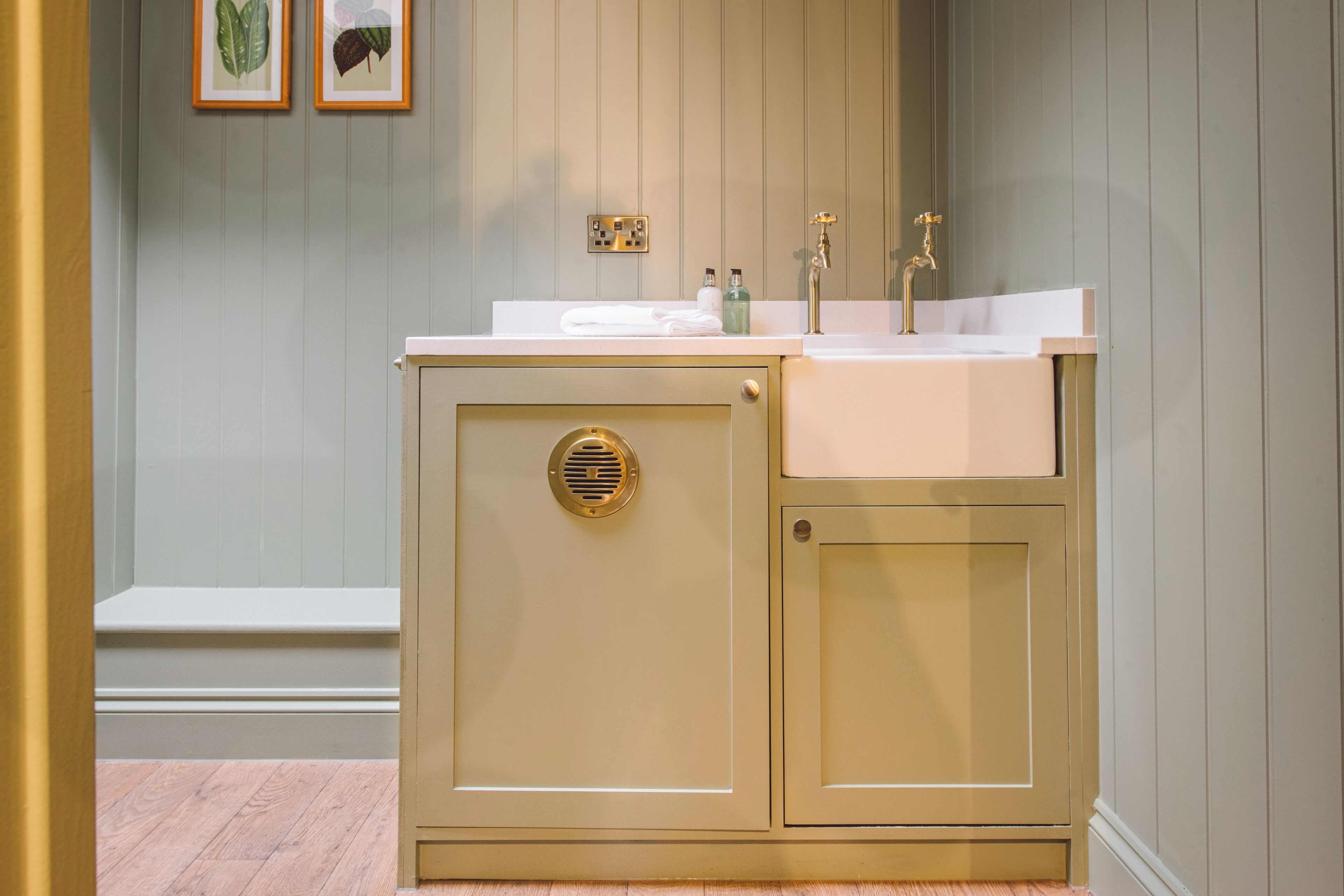 A bathroom with a beige vanity, a white sink, and a gold faucet. There are two framed botanical prints on the wall.