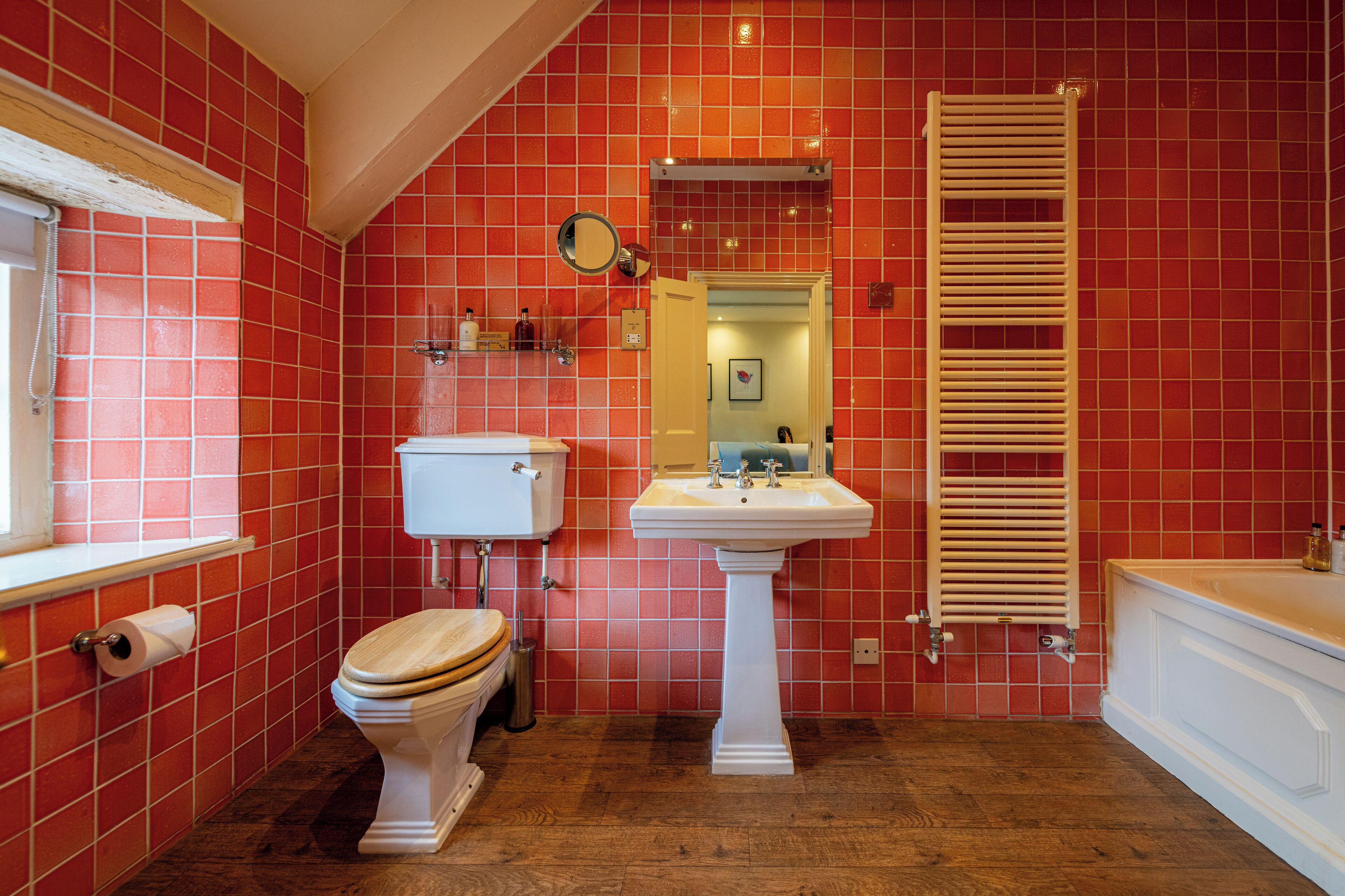 A bathroom with red tiled walls, a white toilet, a pedestal sink, and a wooden towel rack.