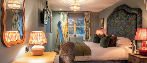 A bedroom with a large bed, a wooden nightstand, a chair, a desk with a lamp, a mirror, and a television on the wall.