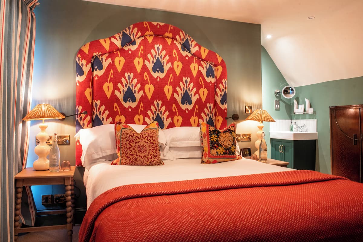 Habitación de hotel con una cama grande, dos mesitas de noche con lámparas, un espejo y un baño con lavamanos y armarios.