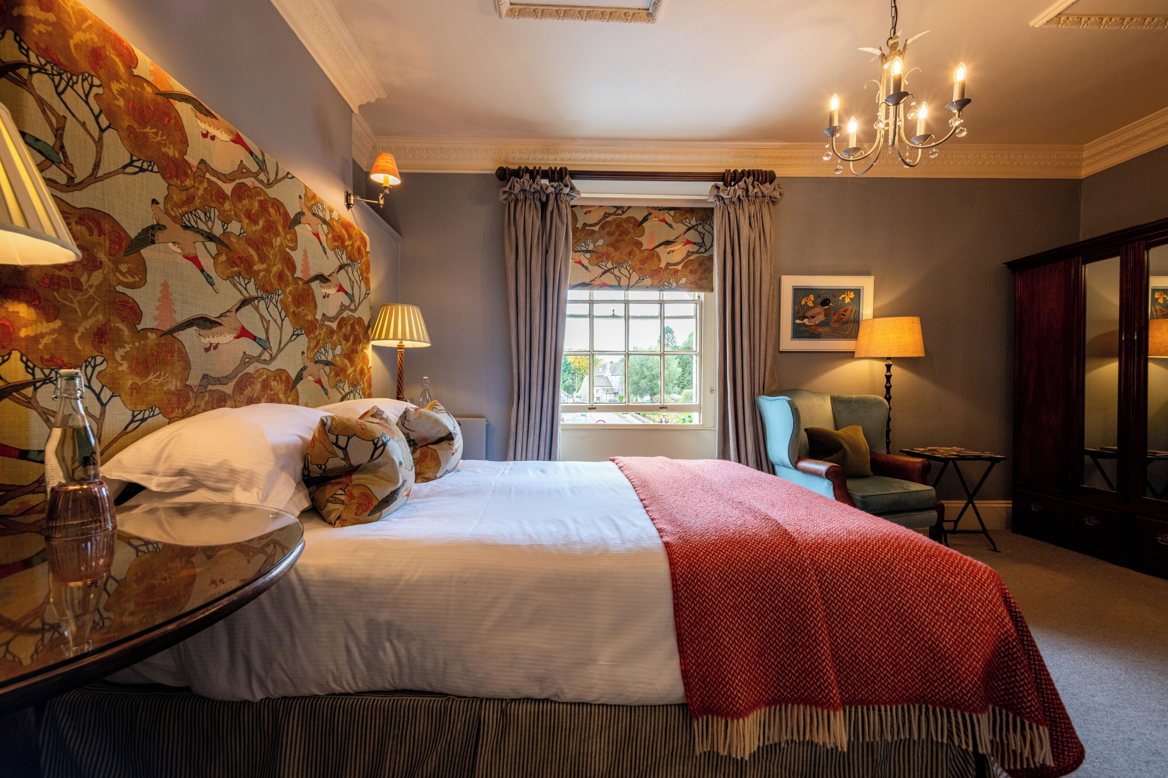 Une chambre d’hôtel avec un grand lit, un coin salon avec un fauteuil, une table avec une lampe et une fenêtre avec des rideaux.