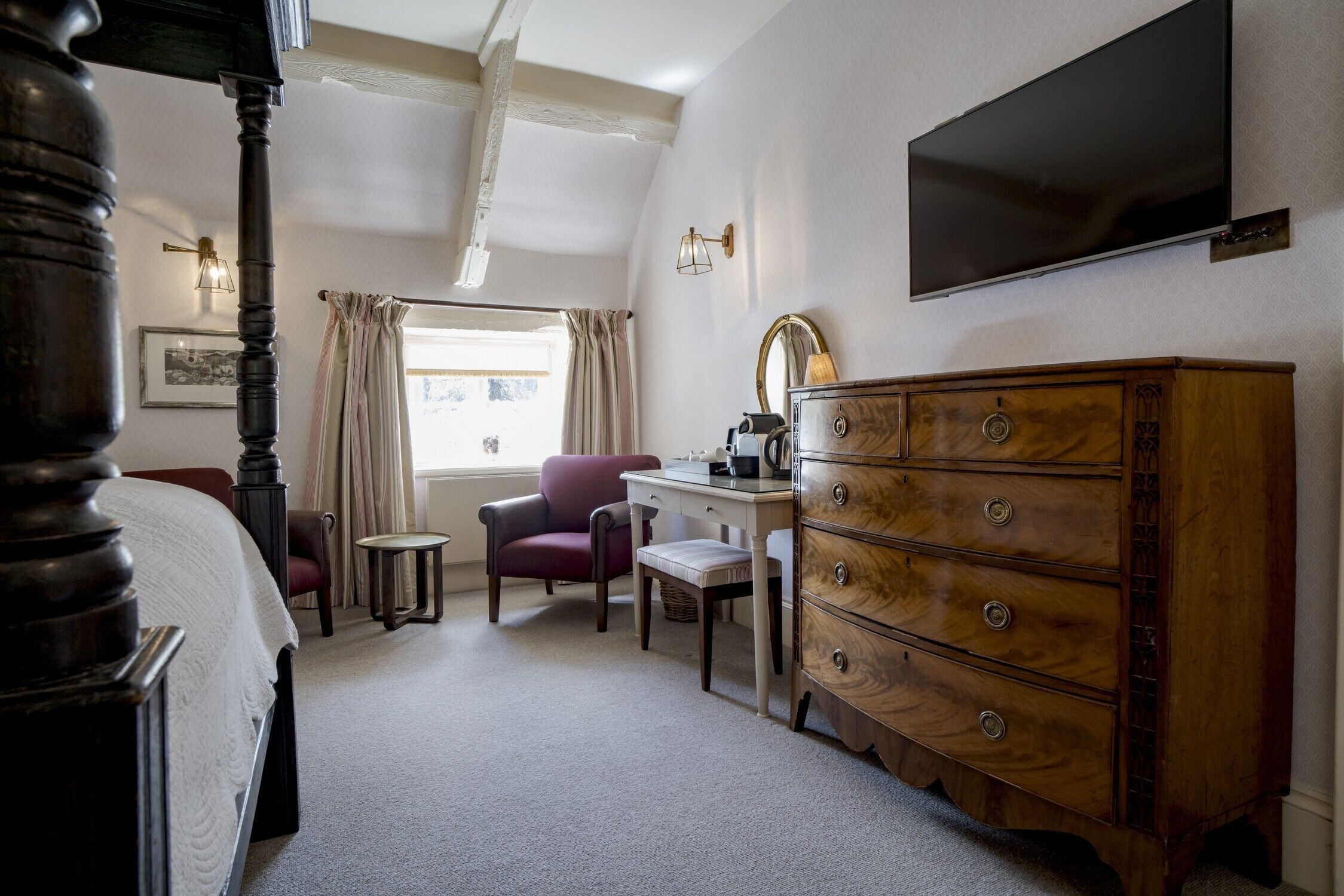 Een slaapkamer met een hemelbed, een houten dressoir, een bureau met stoel, een televisie en een raam met gordijnen.
