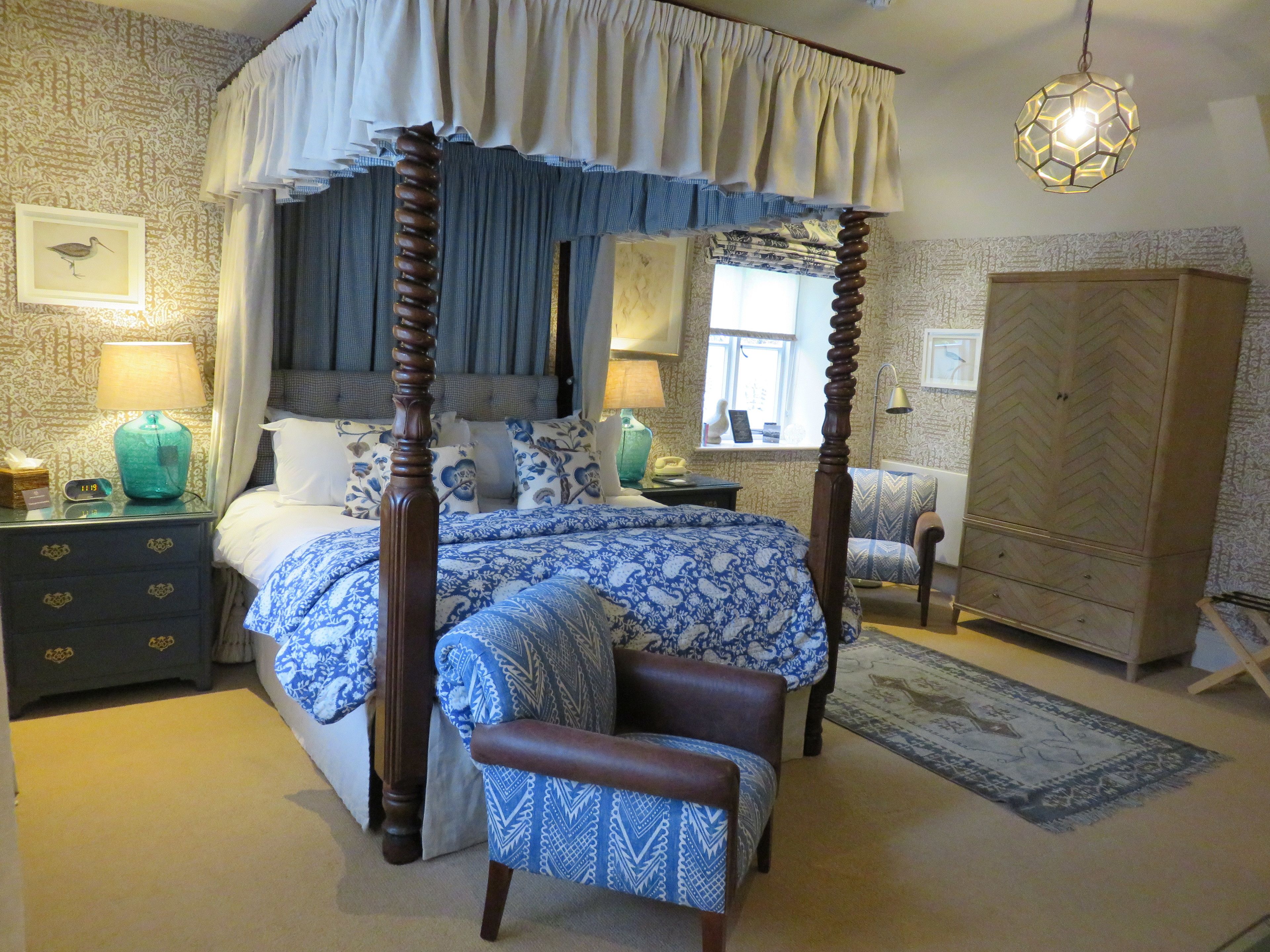 A four-poster canopy bed with a blue and white patterned bedspread, a wooden nightstand with a lamp, a bedside table with a telephone, and a large herringbone-patterned wardrobe.