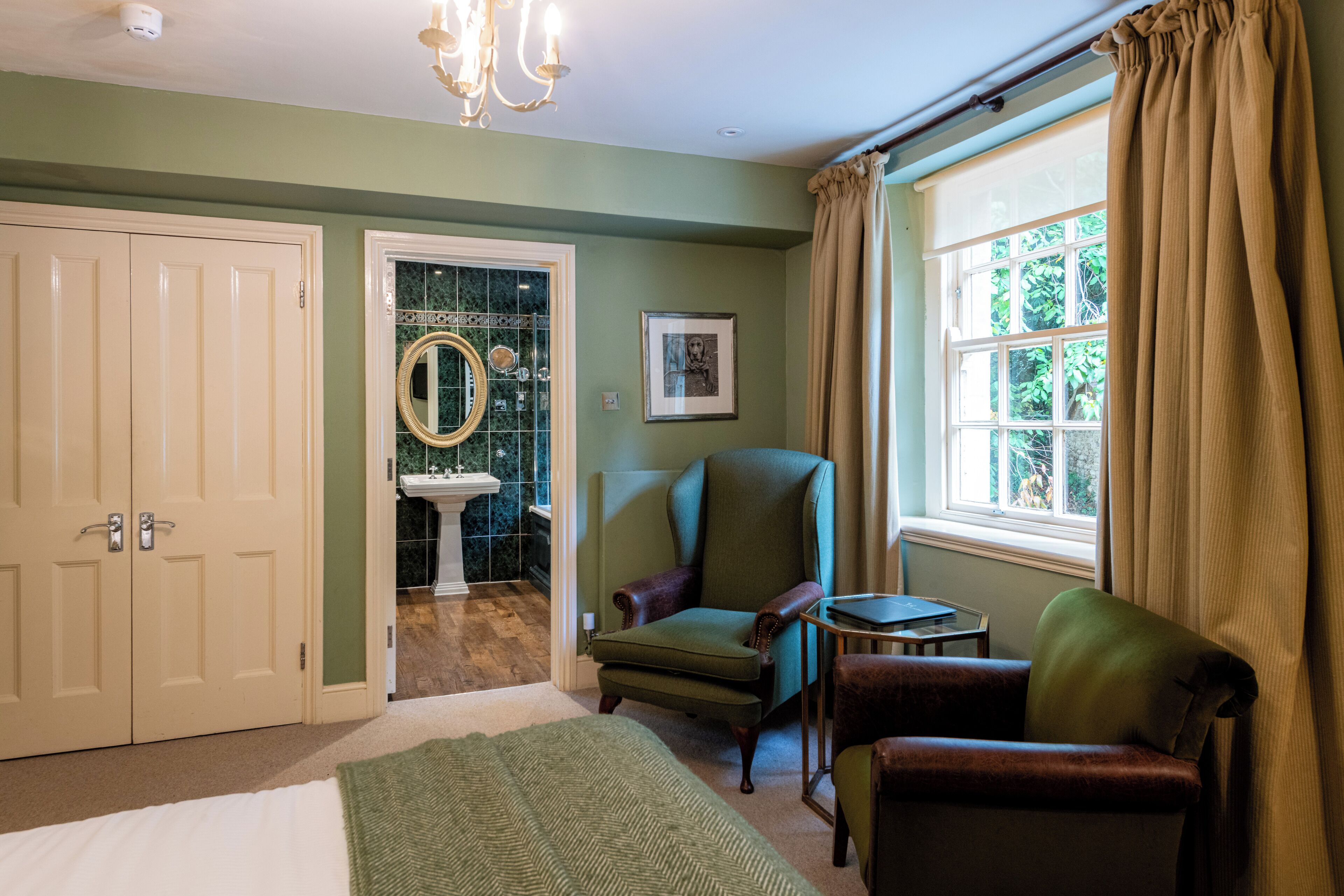 A bedroom with a bed, a chair, a small table, a mirror, and a window with curtains.