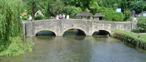A stone bridge over a calm river with ducks swimming, surrounded by lush greenery and a few people walking.