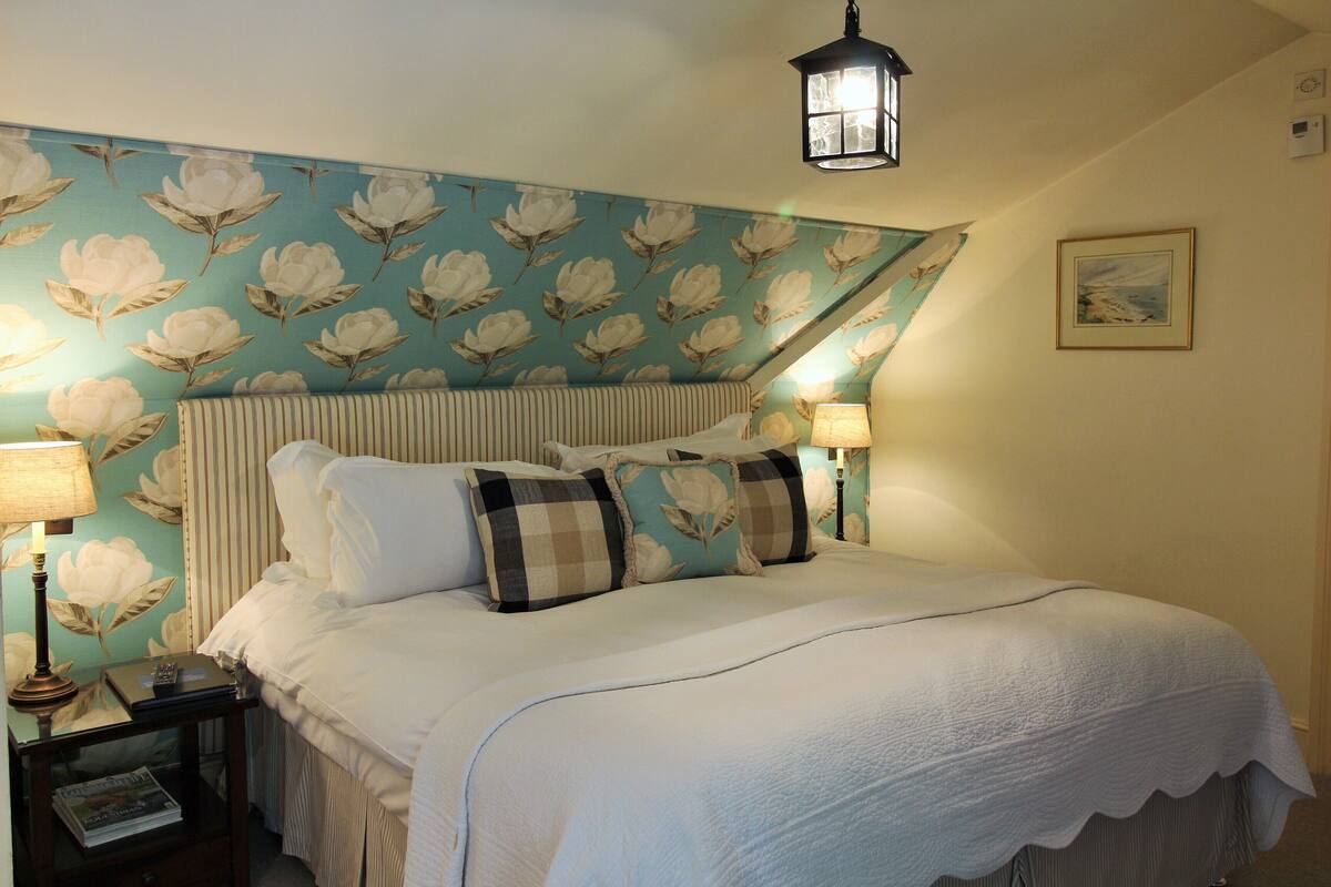 Una cama bien tendida con cabecera floral, dos lámparas de noche y una mesita con una lámpara y un libro.