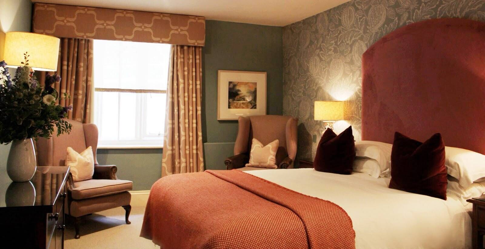 Habitació d'hotel amb un llit gran, dues butaques, un escriptori, un vas amb flors i una finestra amb cortines.