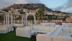 Una terraza en la azotea con muebles de mimbre blanco, un arco decorativo y vistas a un pueblo histórico con edificios en una colina.