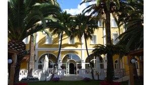 Un edificio amarillo con balcones blancos y arcos, rodeado de palmeras y jardines floridos.