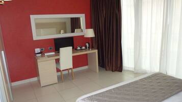 Quarto de hotel com cama, escrivaninha, cadeira, espelho grande, televisão e janela com cortinas.