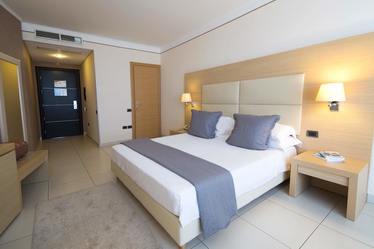 Una habitación de hotel con una cama grande, mesitas de noche, una puerta y una lámpara fijada a la pared.
