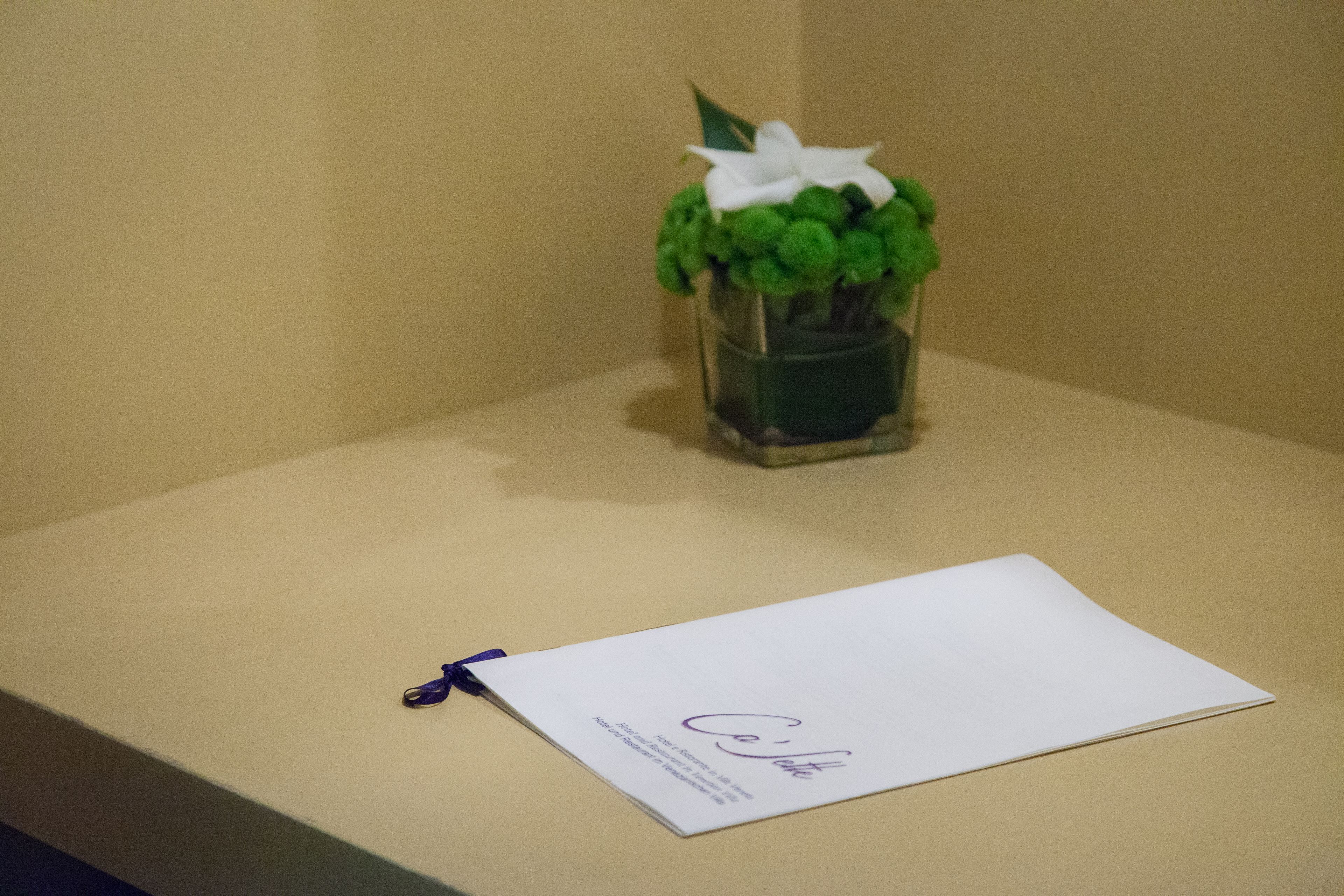 A white card with a signature on it, placed on a flat surface next to a small glass vase with a green plant.