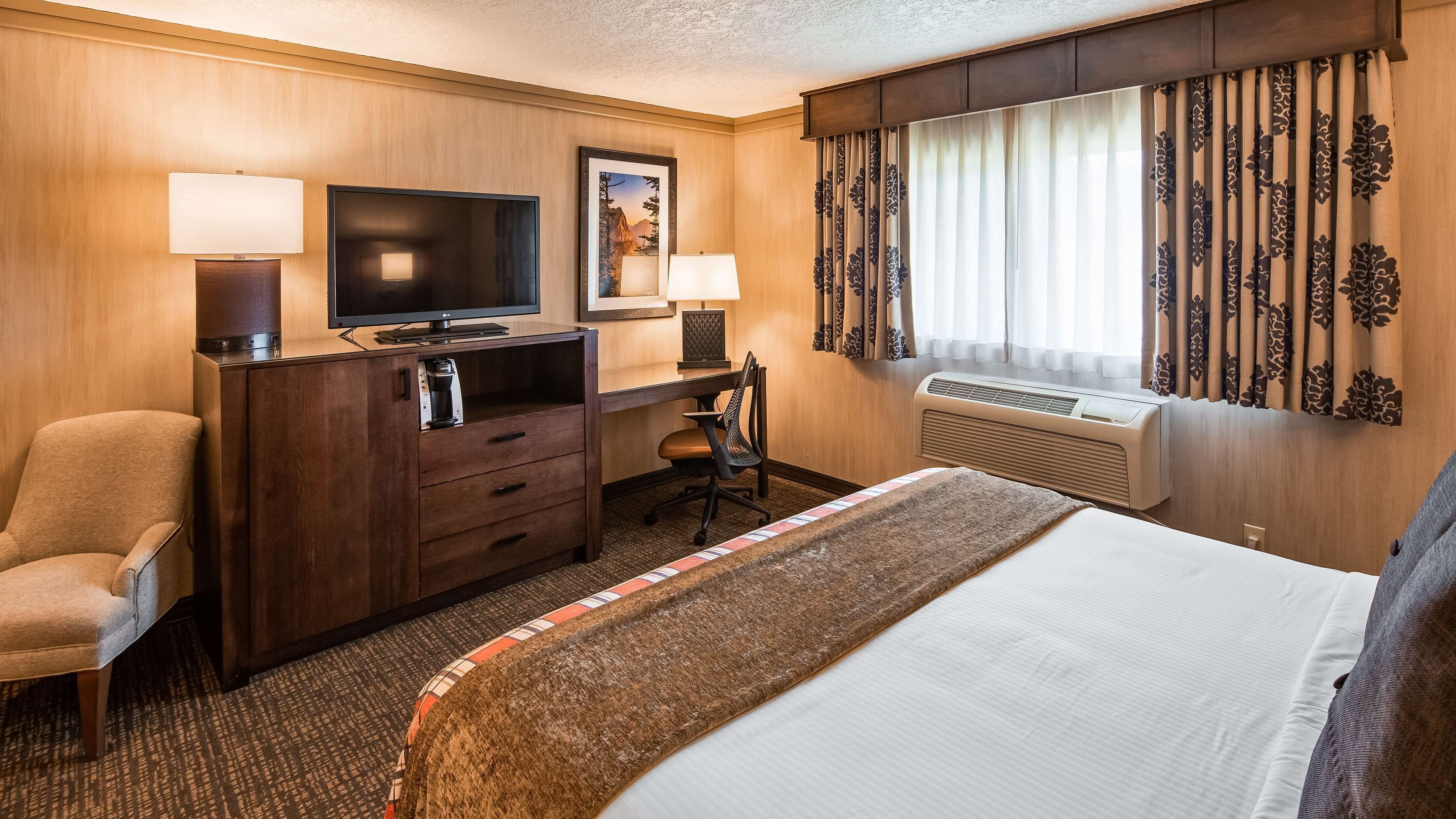 Ein Hotelzimmer mit einem Bett, einem Schreibtisch mit Computer, einem Sessel, einem Fernseher und einem Fenster mit Vorhängen.