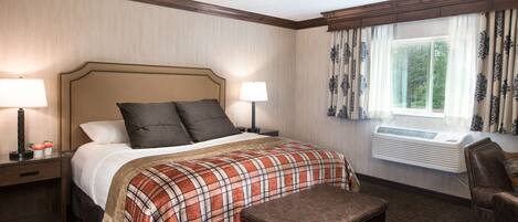 A hotel room with a bed, two bedside lamps, a window with curtains, an air conditioning unit, a chair, and a small table.