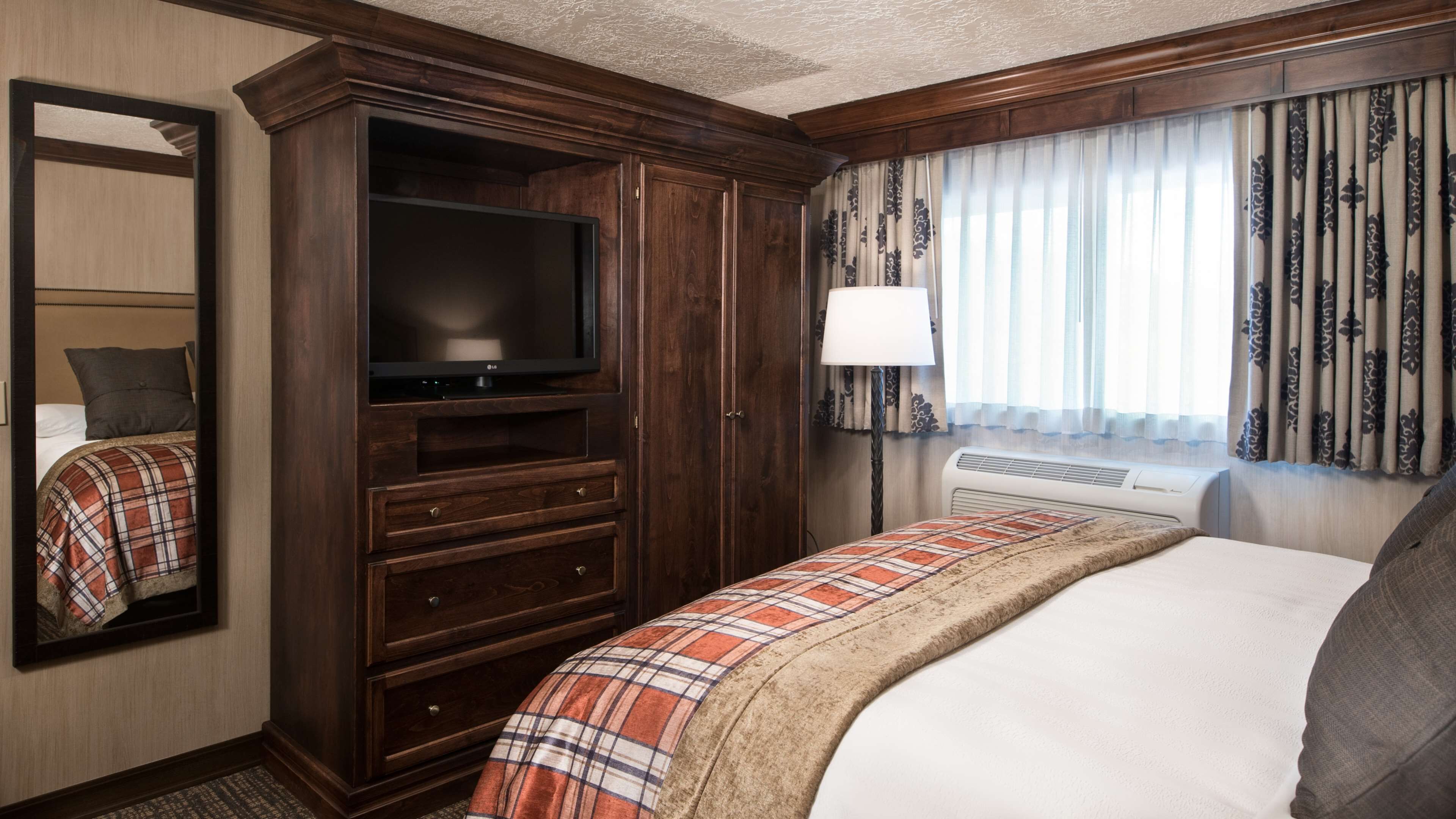 Ein Hotelzimmer mit einem großen Bett, einem Fernseher auf einer Holzkommode, einem Spiegel, einem Fenster mit Vorhängen und einem Nachttisch mit Lampe.