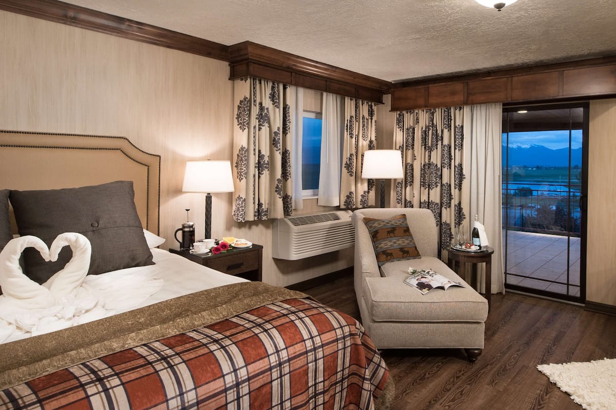 A hotel room with a bed, a sofa, a small table with a lamp, a window with curtains, and a view of the outdoors.