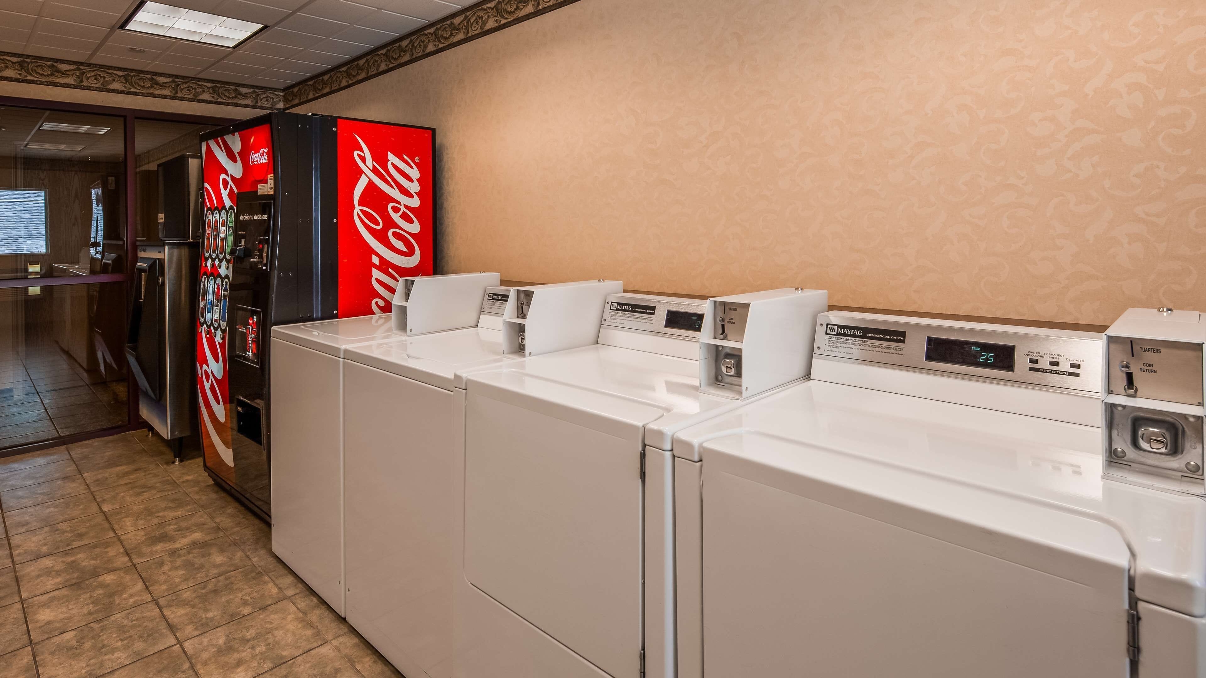 Ein Hotel-Waschraum mit vier weißen Waschmaschinen und Trocknern, einer Coca-Cola-Getränkeautomaten und einem Maytag-Trockner für den gewerblichen Gebrauch.