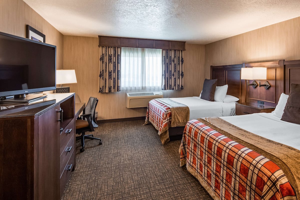 A hotel room with two beds, a desk with a TV, a chair, a window with curtains, and an air conditioning unit.