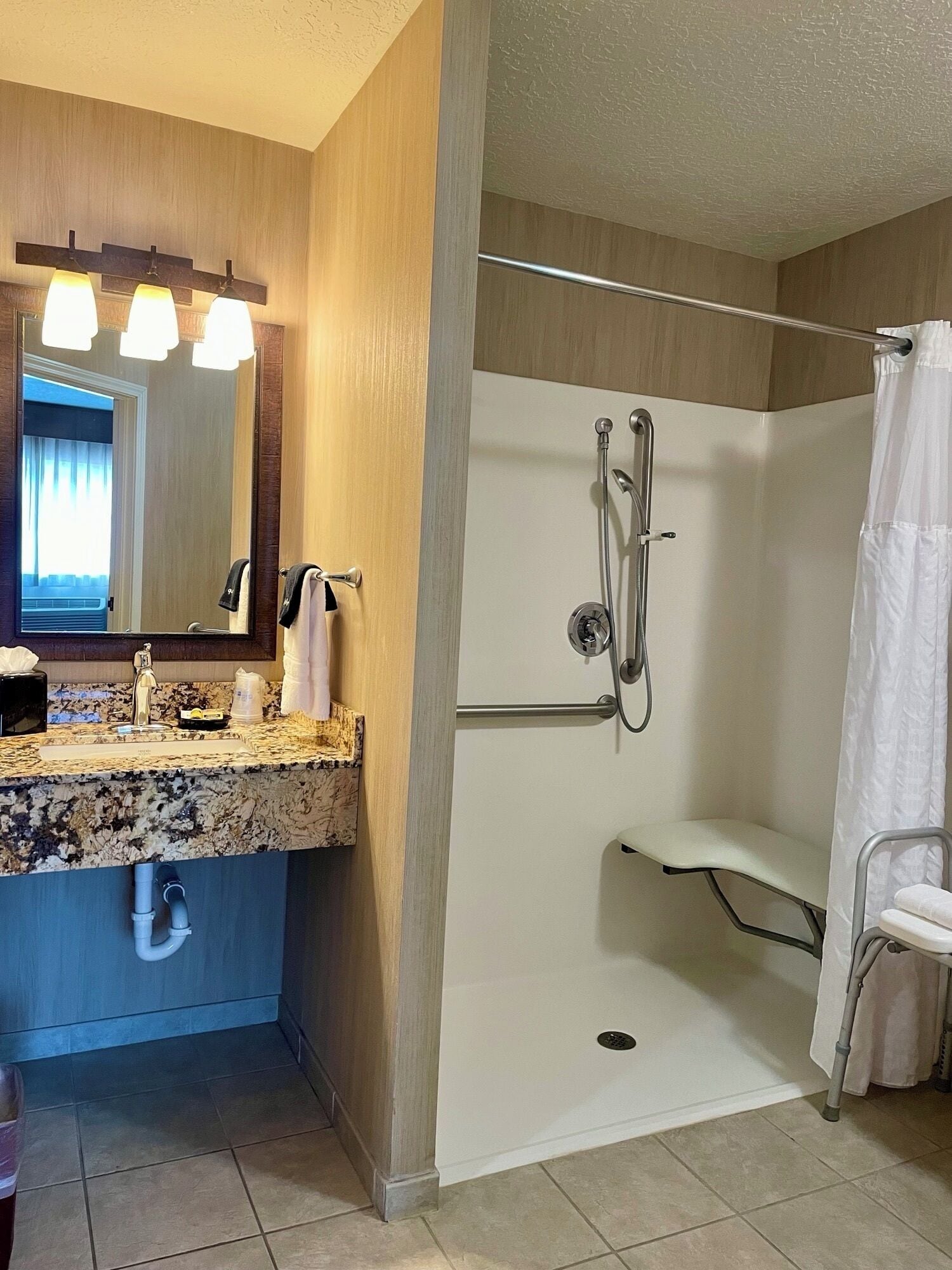 Un baño con encimera de granito, un lavabo, un espejo, ducha con puerta de cristal y una silla blanca.
