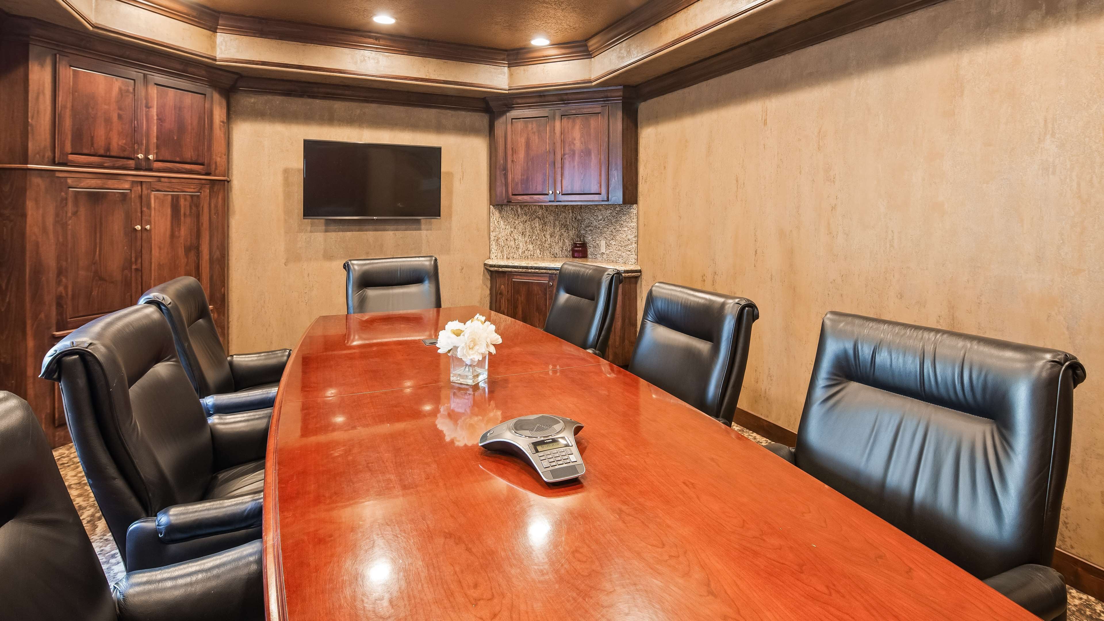 A conference room with a long wooden table, black leather chairs, a small vase with flowers, and a speaker.